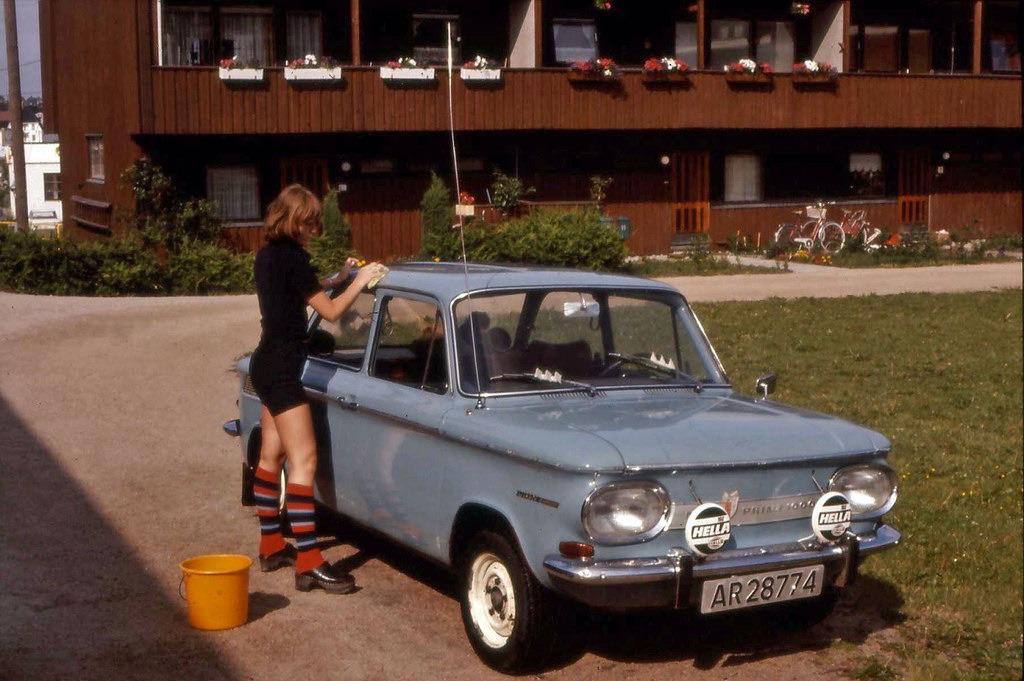 A person washing a light blue NSU Prinz car parked in front of a house.