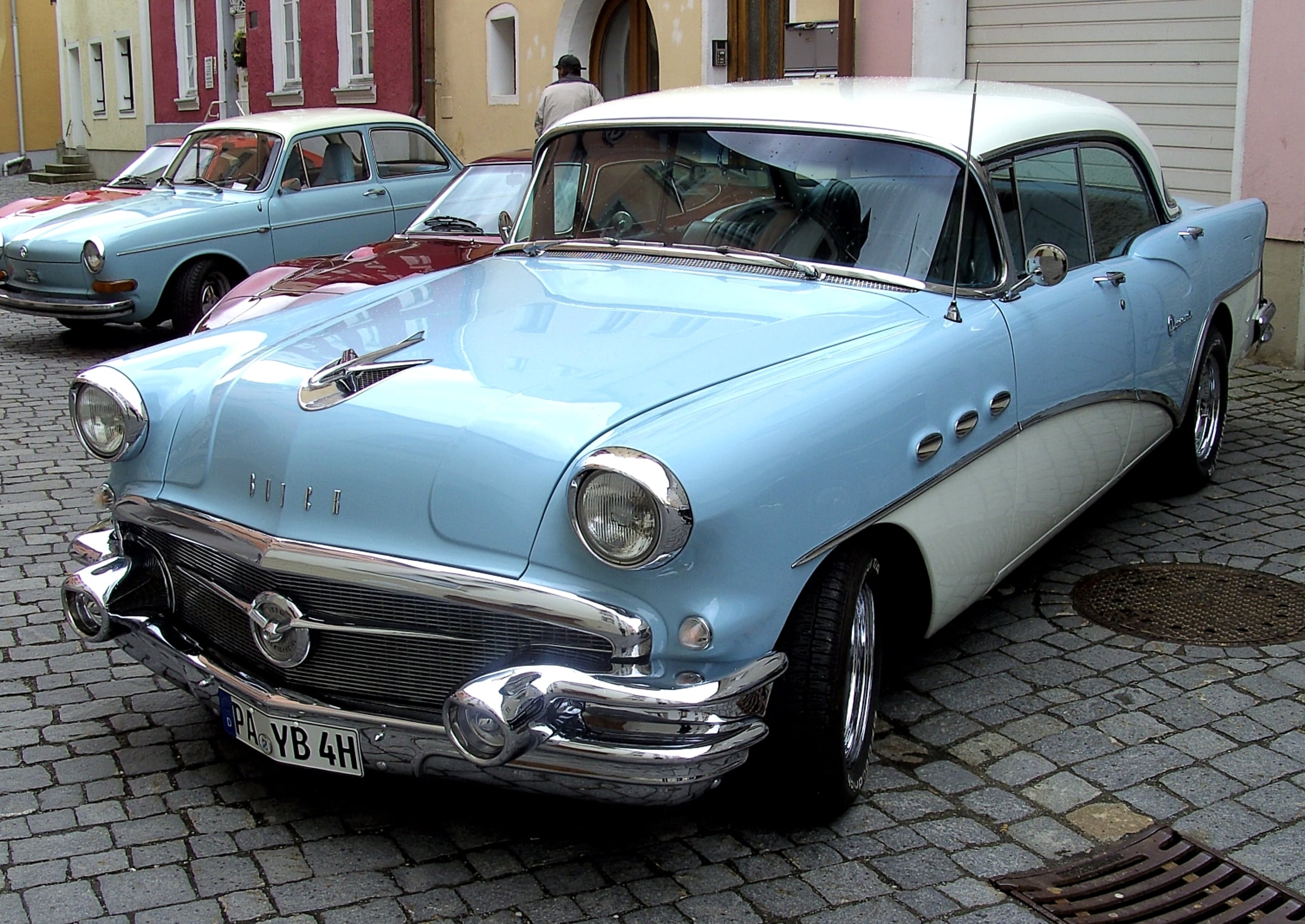 A vintage light blue and white Buick parked on a cobblestone street with a light blue Volkswagen parked behind it.