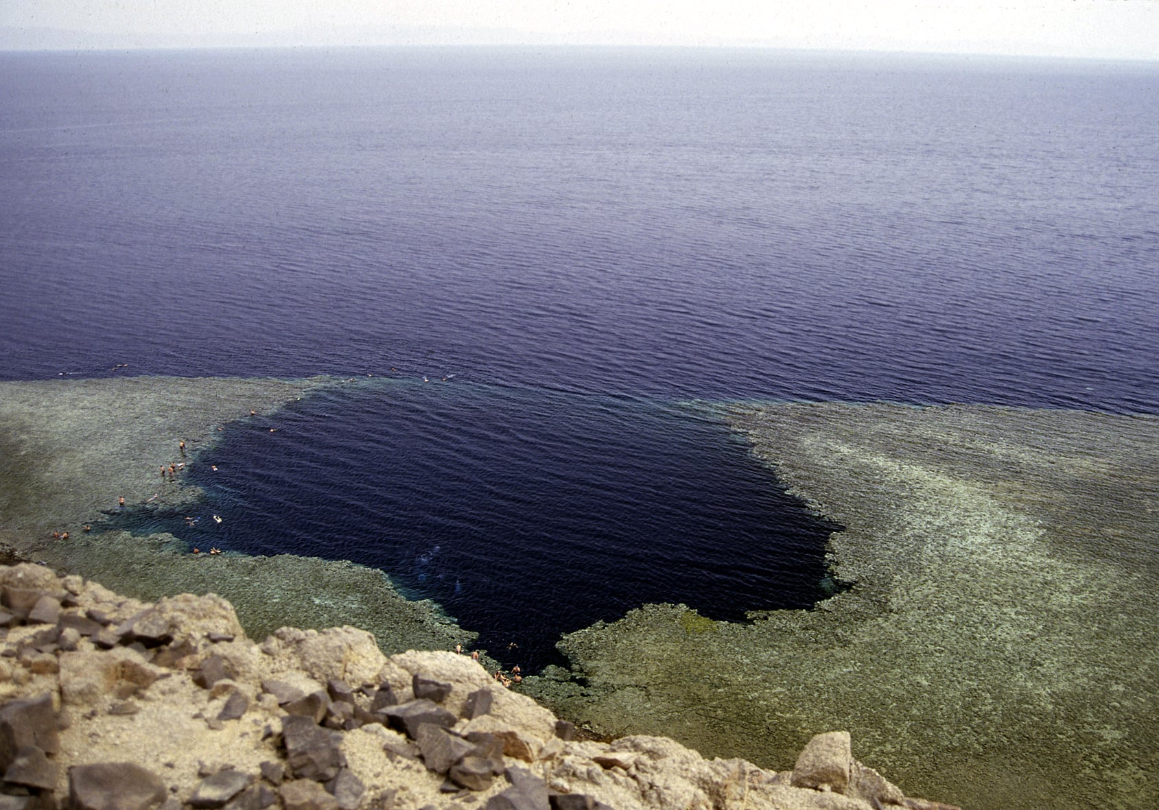 A large, deep-blue circular sinkhole surrounded by a shallow, light-colored coral reef in the middle of a vast sea, viewed from a rocky cliff.