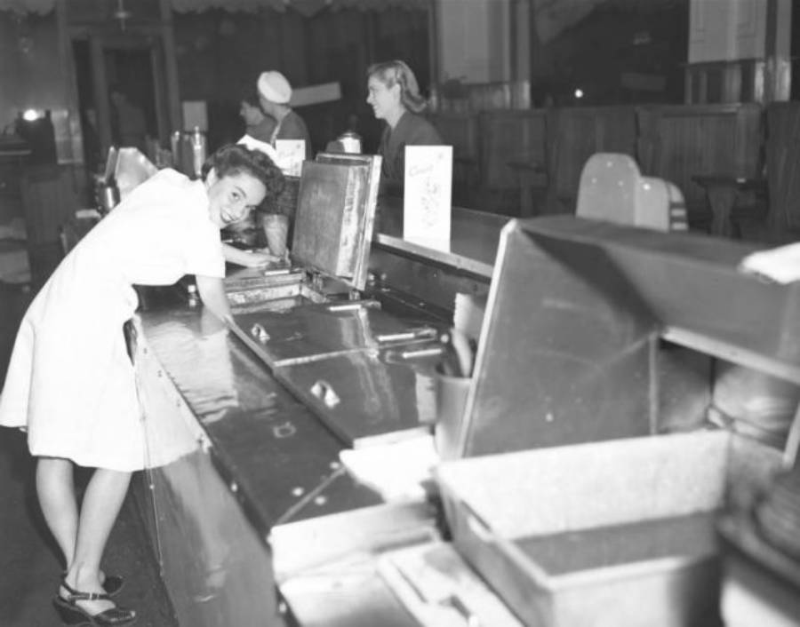 A smiling woman in a white uniform stands behind a lunch counter, looking toward the camera.