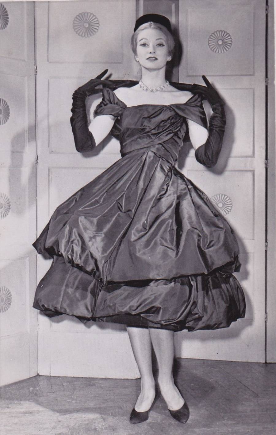 A woman posing in an off-the-shoulder, tea-length evening gown with long black gloves and a small pillbox hat.