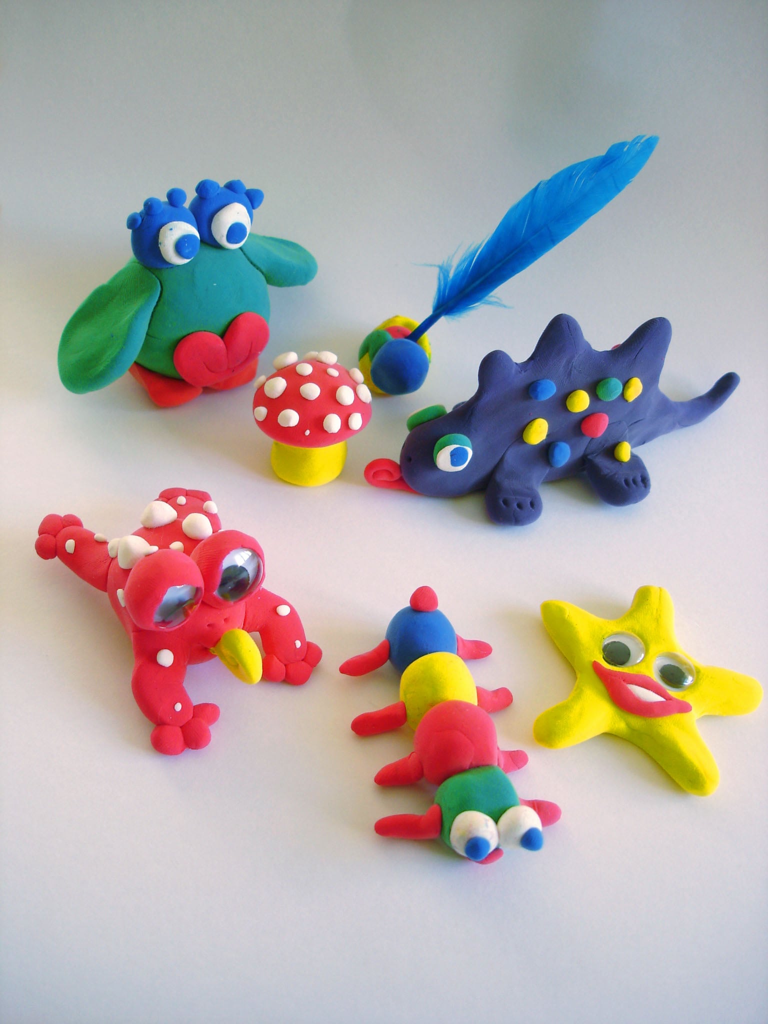 A collection of handmade modeling clay creatures, including a red frog, a blue stegosaurus with a feather, a multi-colored caterpillar, a yellow star with a face, a green owl-like figure, and a red mushroom.