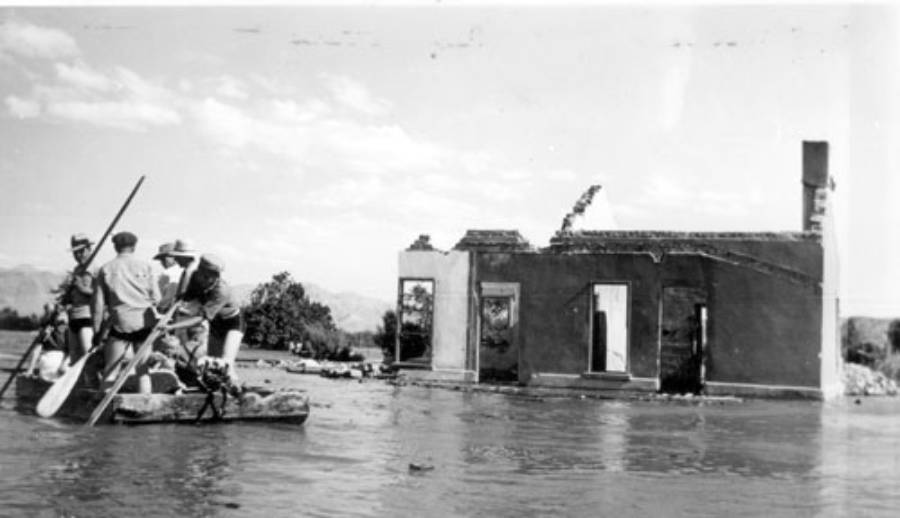 Four people in a small boat navigate through floodwaters past a ruined, partially submerged stone building.