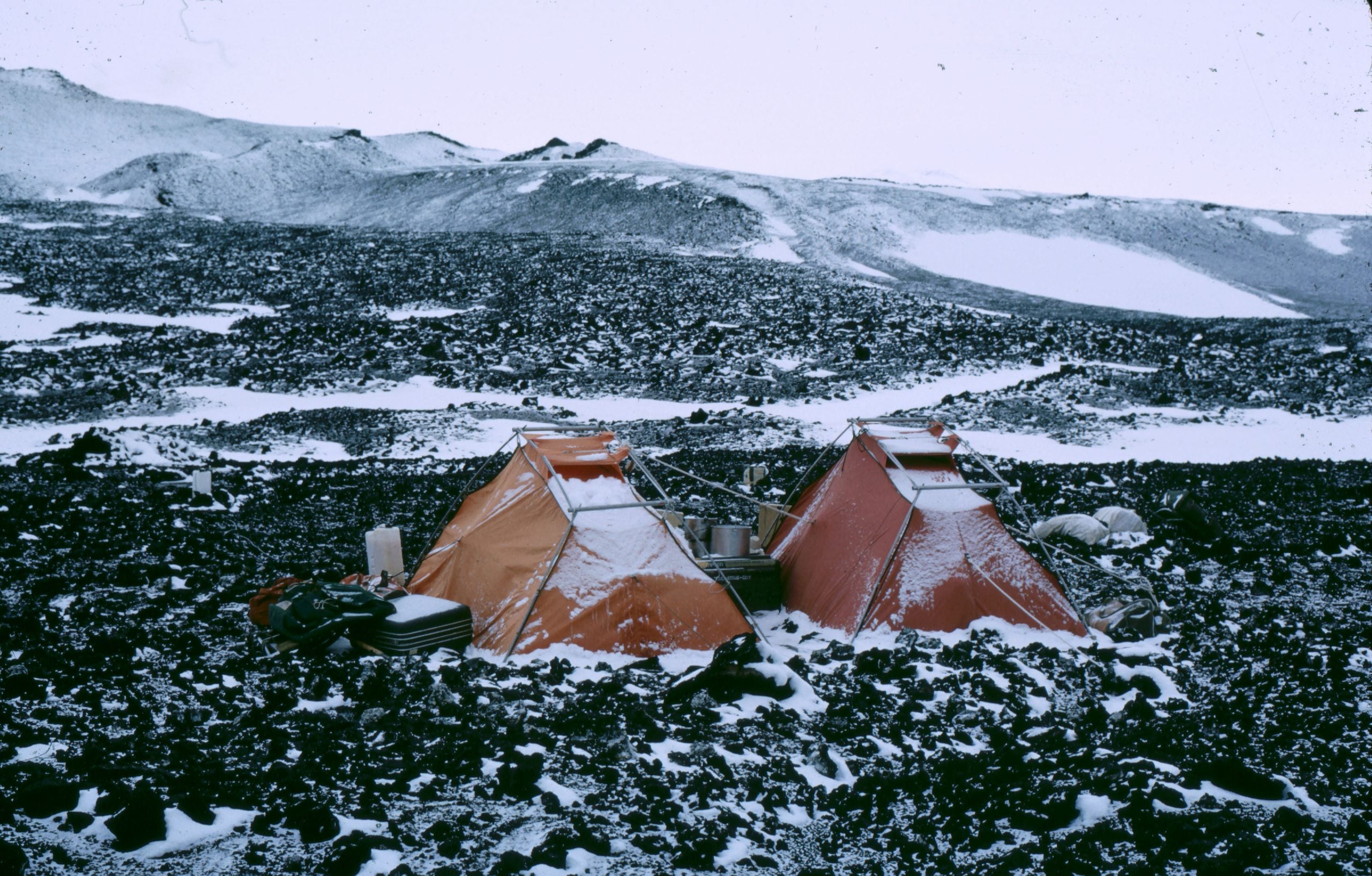 Two orange tents set up on a rocky, snow-covered volcanic landscape.