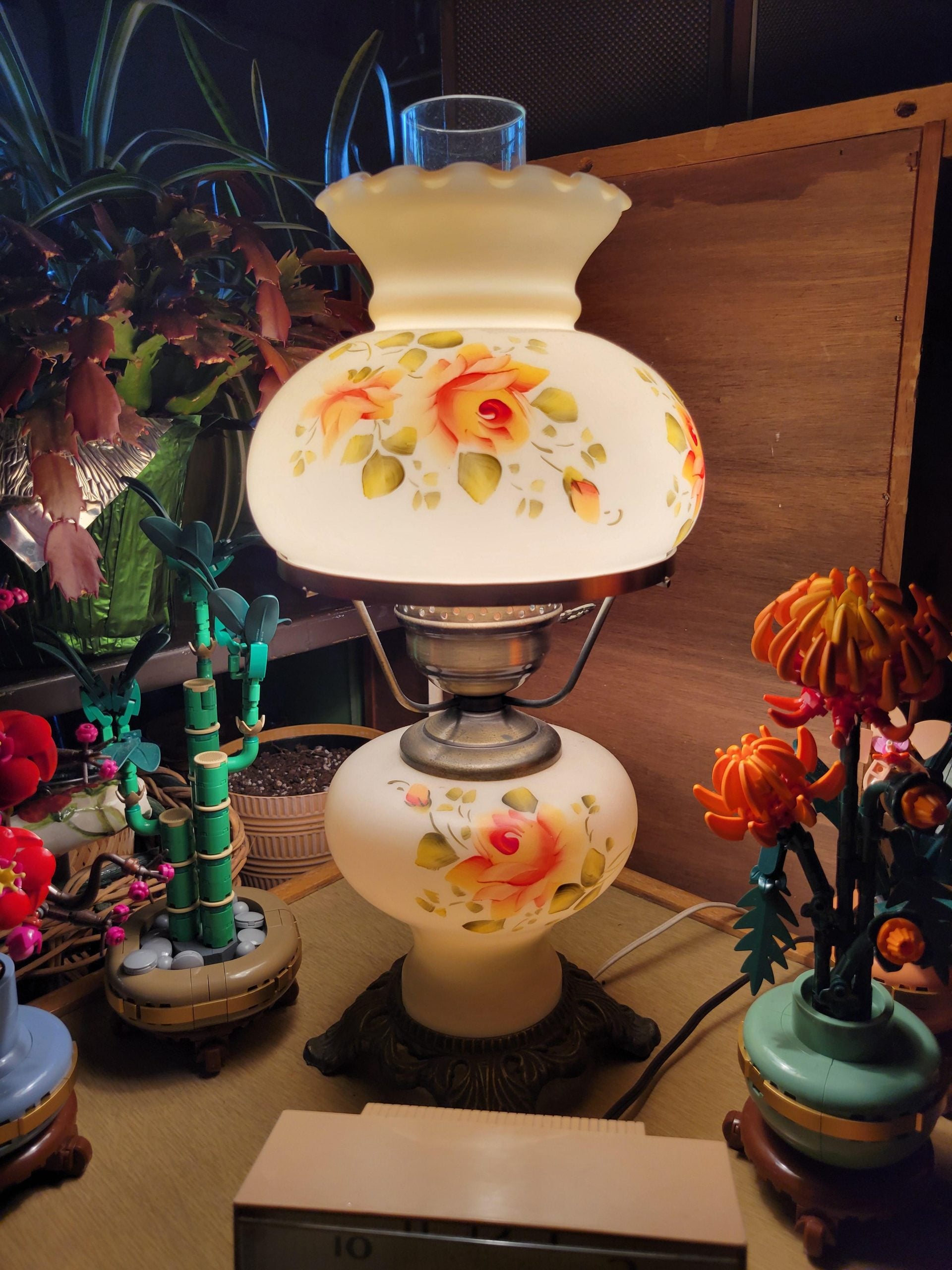 A lighted vintage-style glass lamp with a hand-painted floral design, surrounded by a potted plant and two decorative building-block flower arrangements.