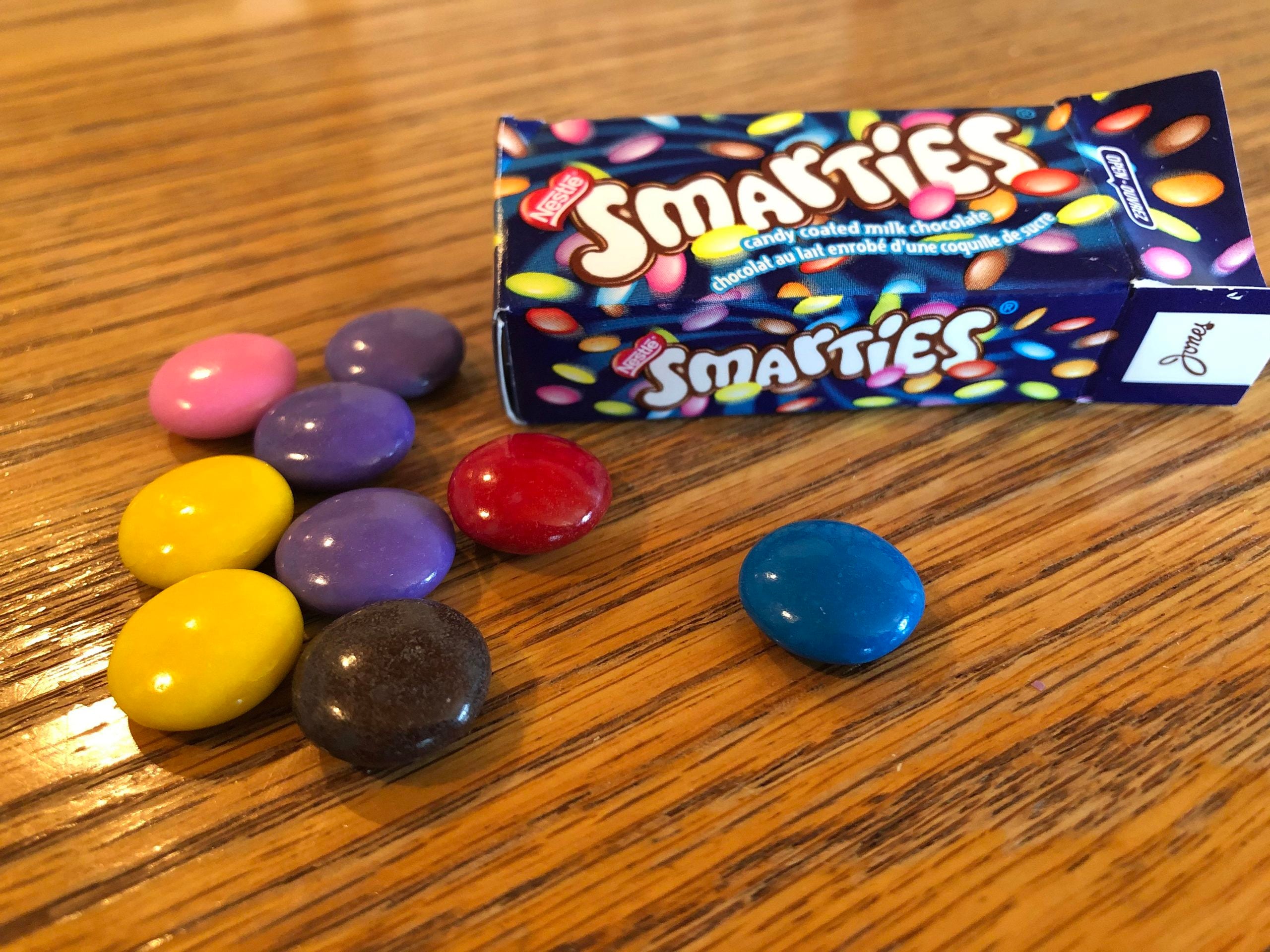 A box of Nestlé Smarties next to a small pile of colorful, candy-coated chocolate pieces on a wooden surface.