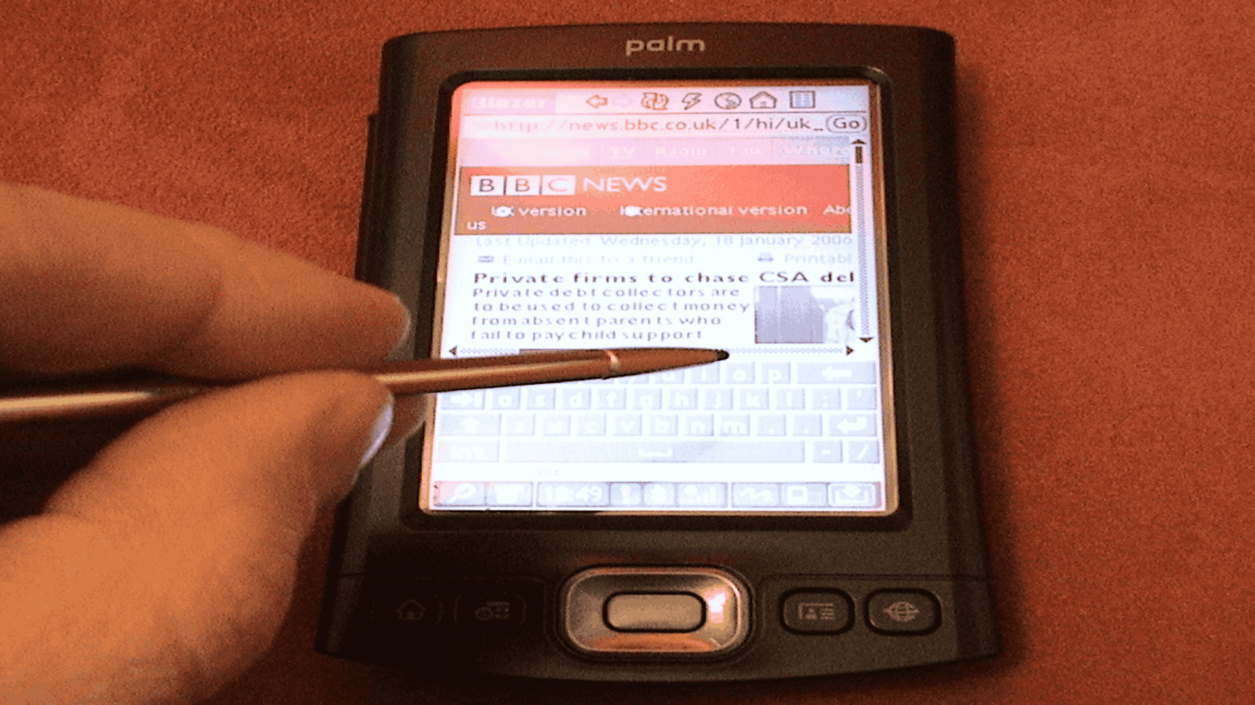 A hand uses a stylus to navigate a BBC News article on a Palm handheld device.