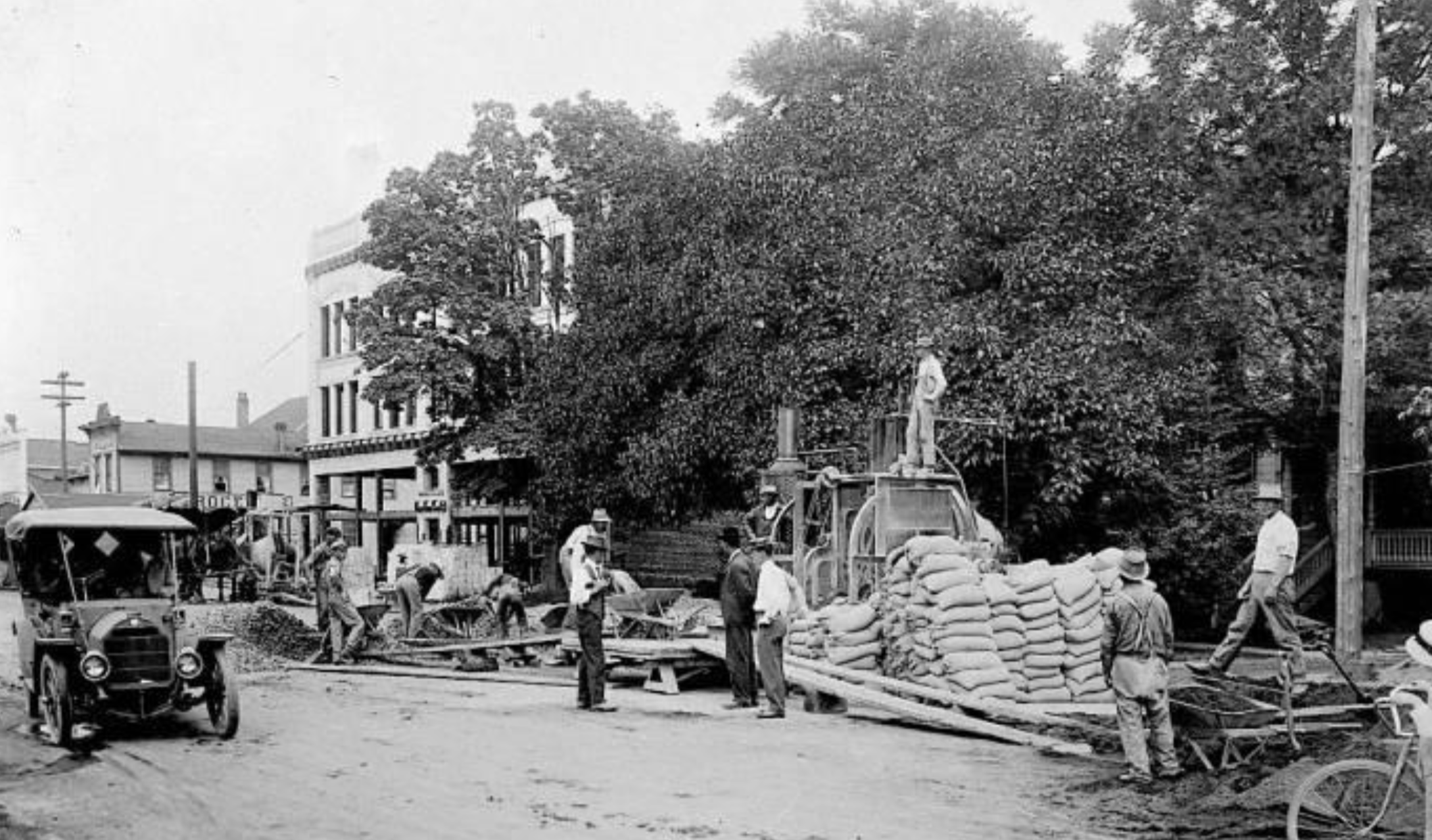 Construction crew working on a dirt road, with an early model automobile parked on the left and a pile of sacks on the right.