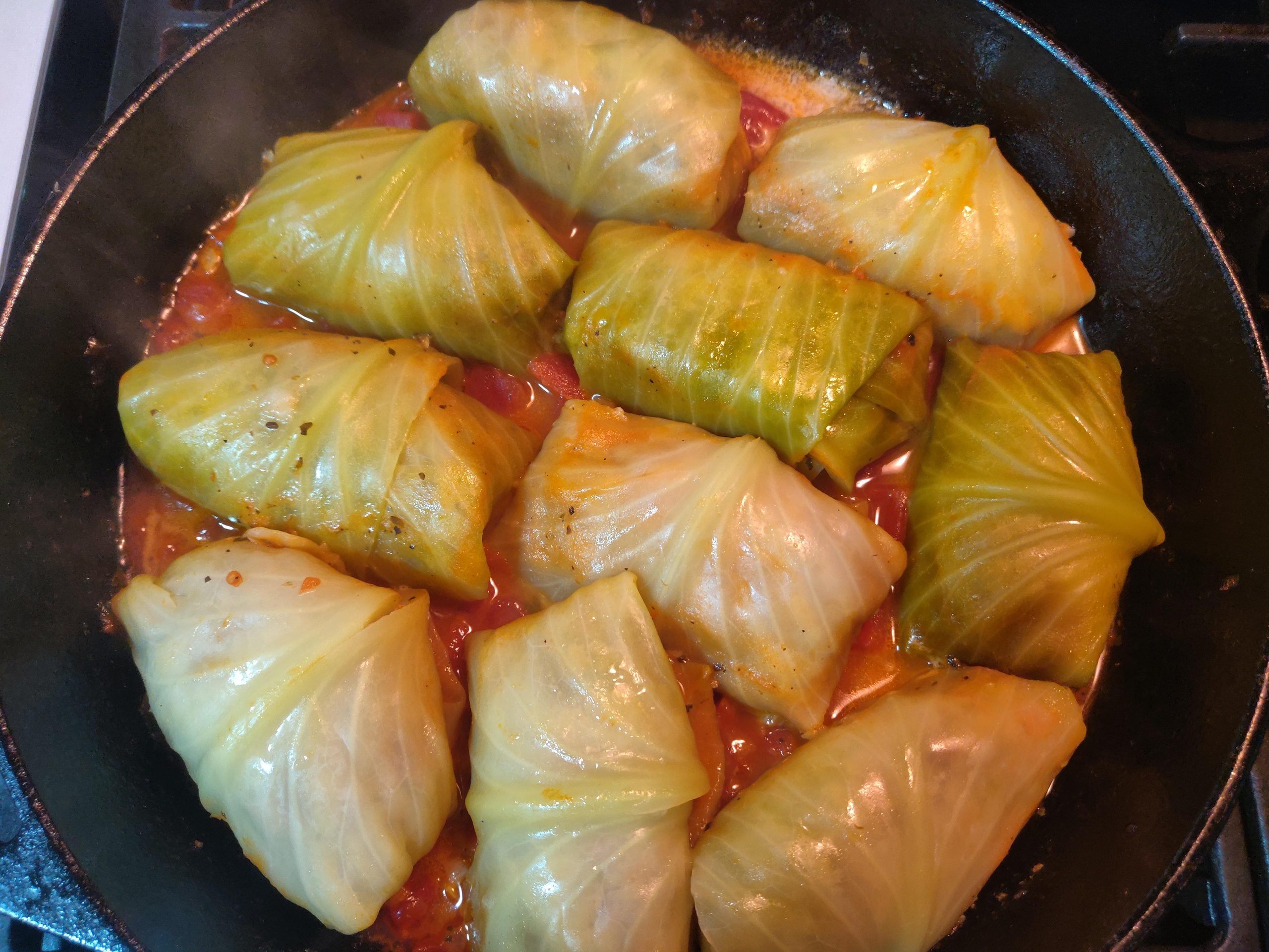 Cabbage rolls filled with meat and rice are cooking in tomato sauce in a black skillet. The rolls are tightly wrapped, and the sauce is visible around the edges.