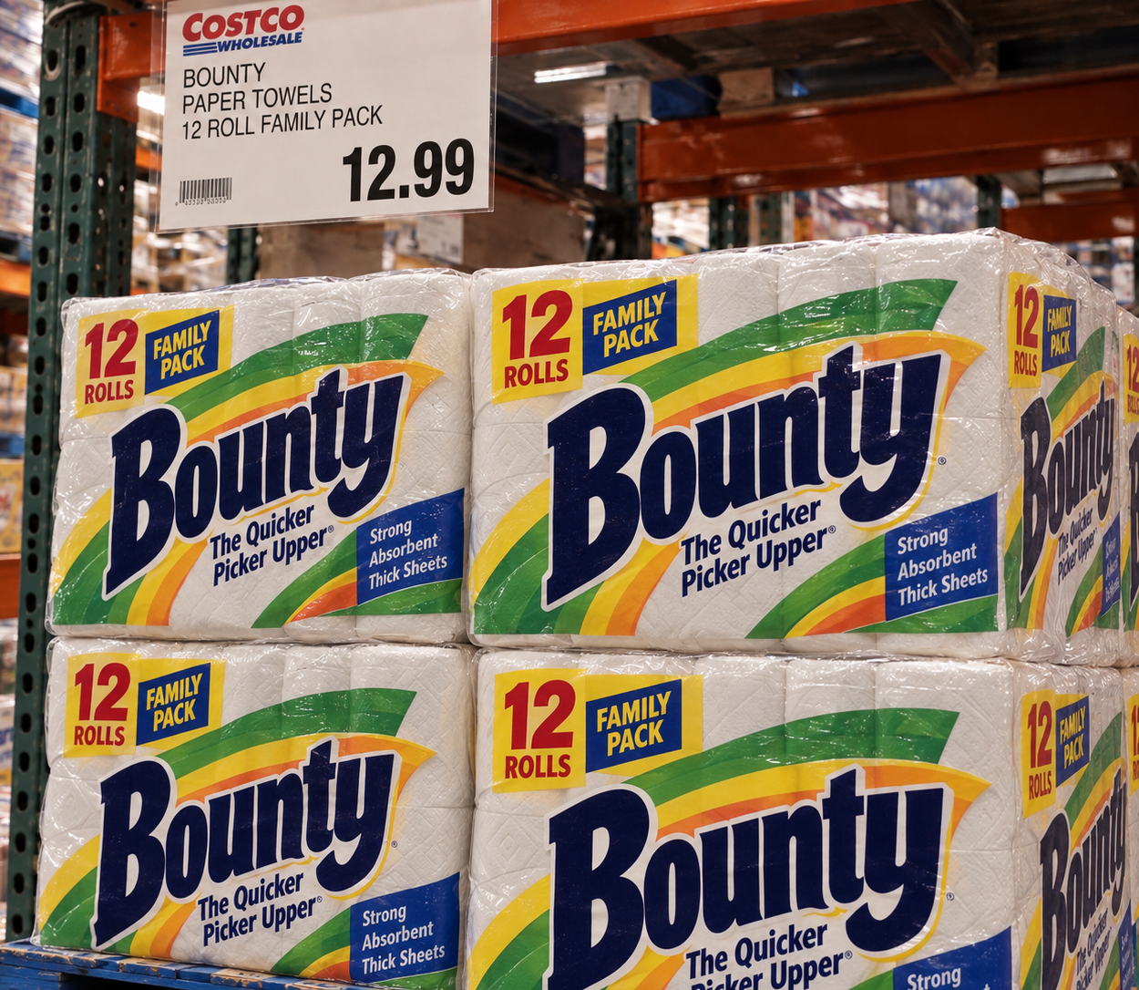 Costco shelf sign for Bounty 12-roll paper towel family packs priced at $12.99, positioned above several stacks of the product.