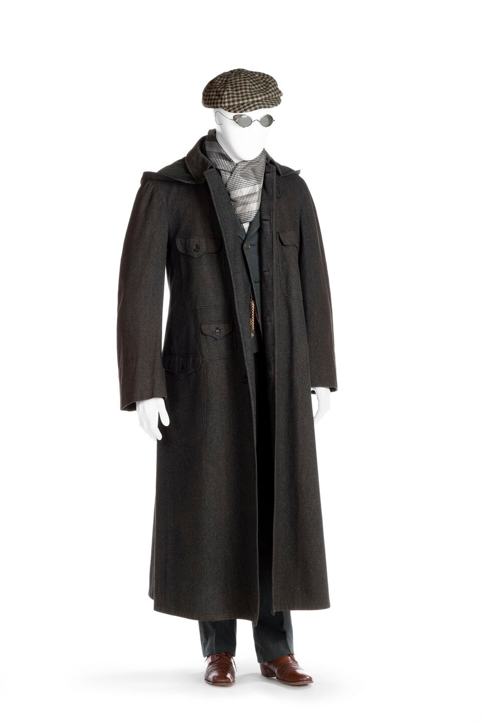 A mannequin wears a long dark trench coat, matching pants, a vest, a striped scarf, brown shoes, round sunglasses, and a plaid newsboy cap against a white background.