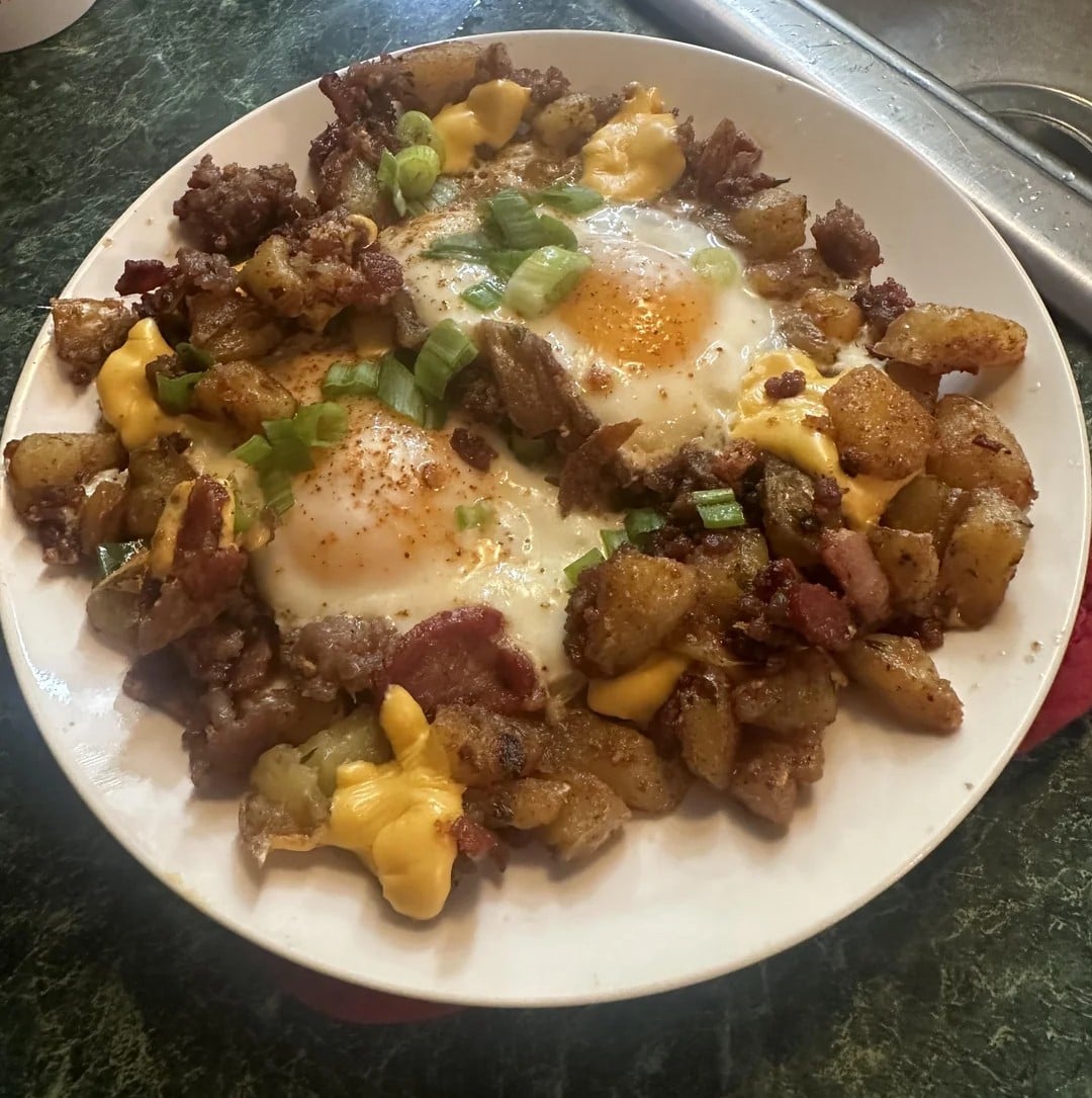 A plate of breakfast hash with crispy potatoes, melted cheese, bacon, sausage, two sunny-side-up eggs, and chopped green onions on top.