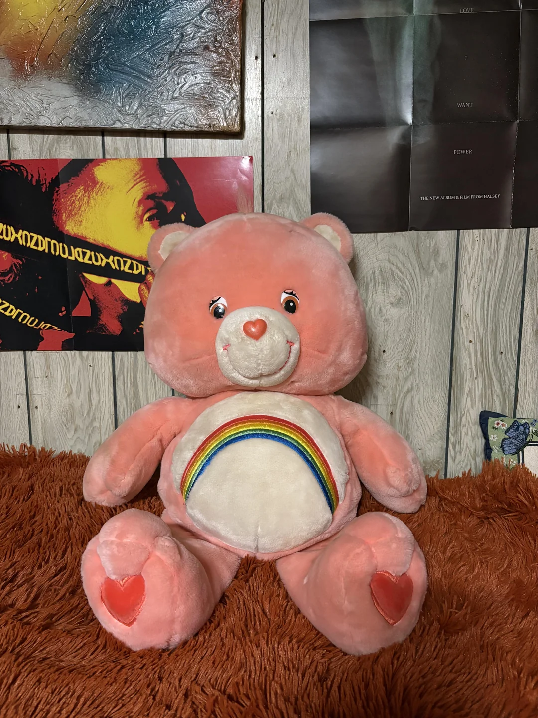 A pink teddy bear with a rainbow on its belly sits on an orange fuzzy surface, against a wall with posters and wood paneling.