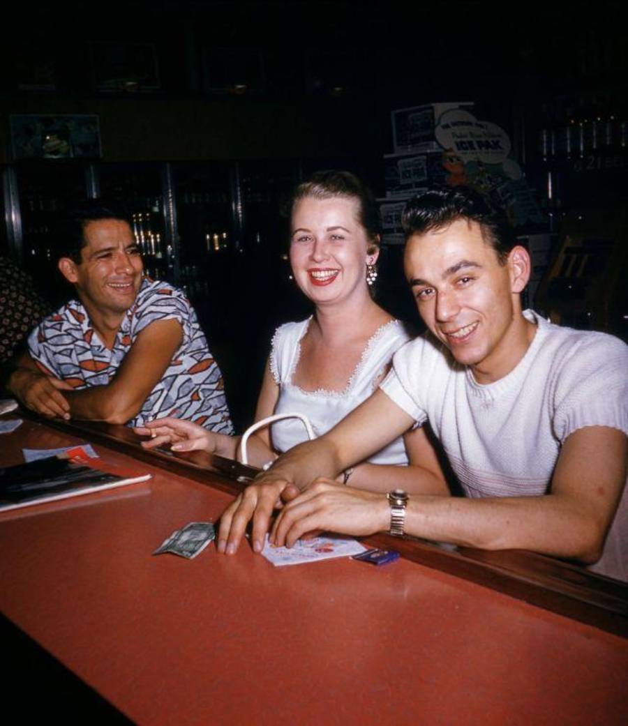 Two men and a woman sit at a bar counter with drinks and money in front of them.