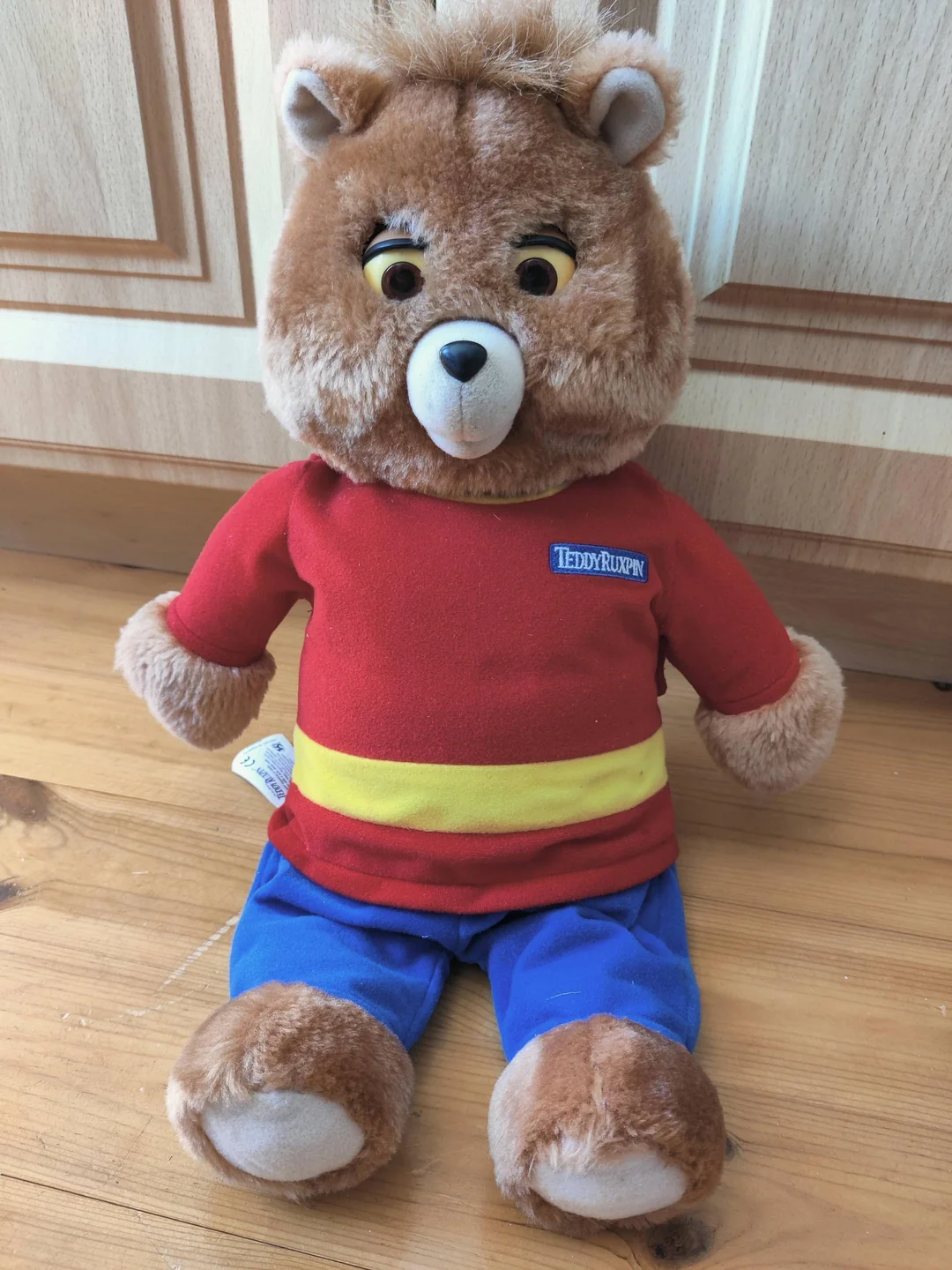 A brown teddy bear plush toy wearing a red shirt with a yellow stripe and blue pants sits on a wooden floor in front of light-colored cabinet doors.