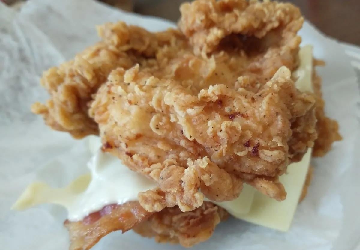 A fried chicken sandwich topped with a slice of cheese and a strip of bacon on parchment paper.