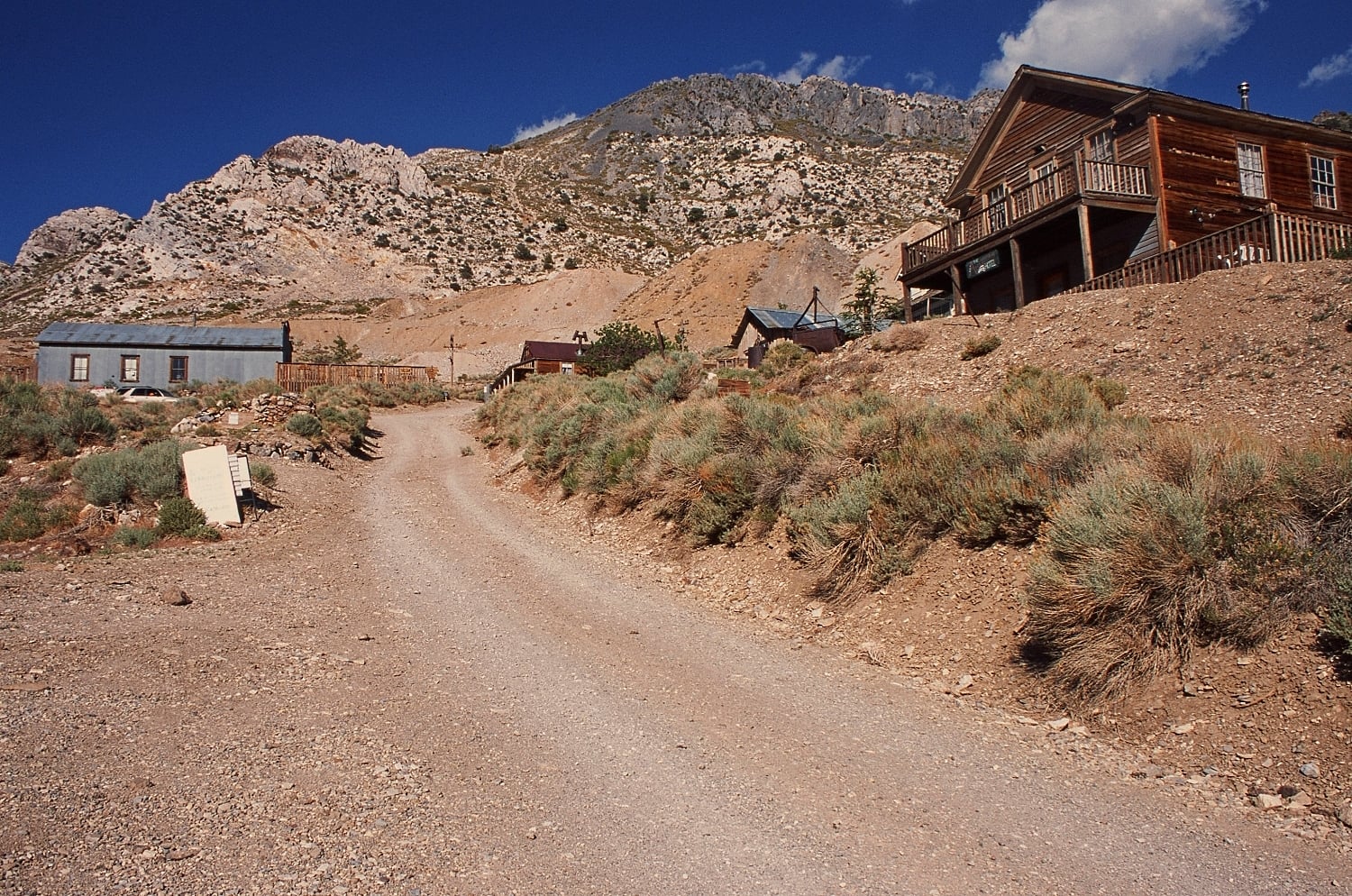 A dirt road leads through a desert ghost town toward a rocky mountain under a clear blue sky.