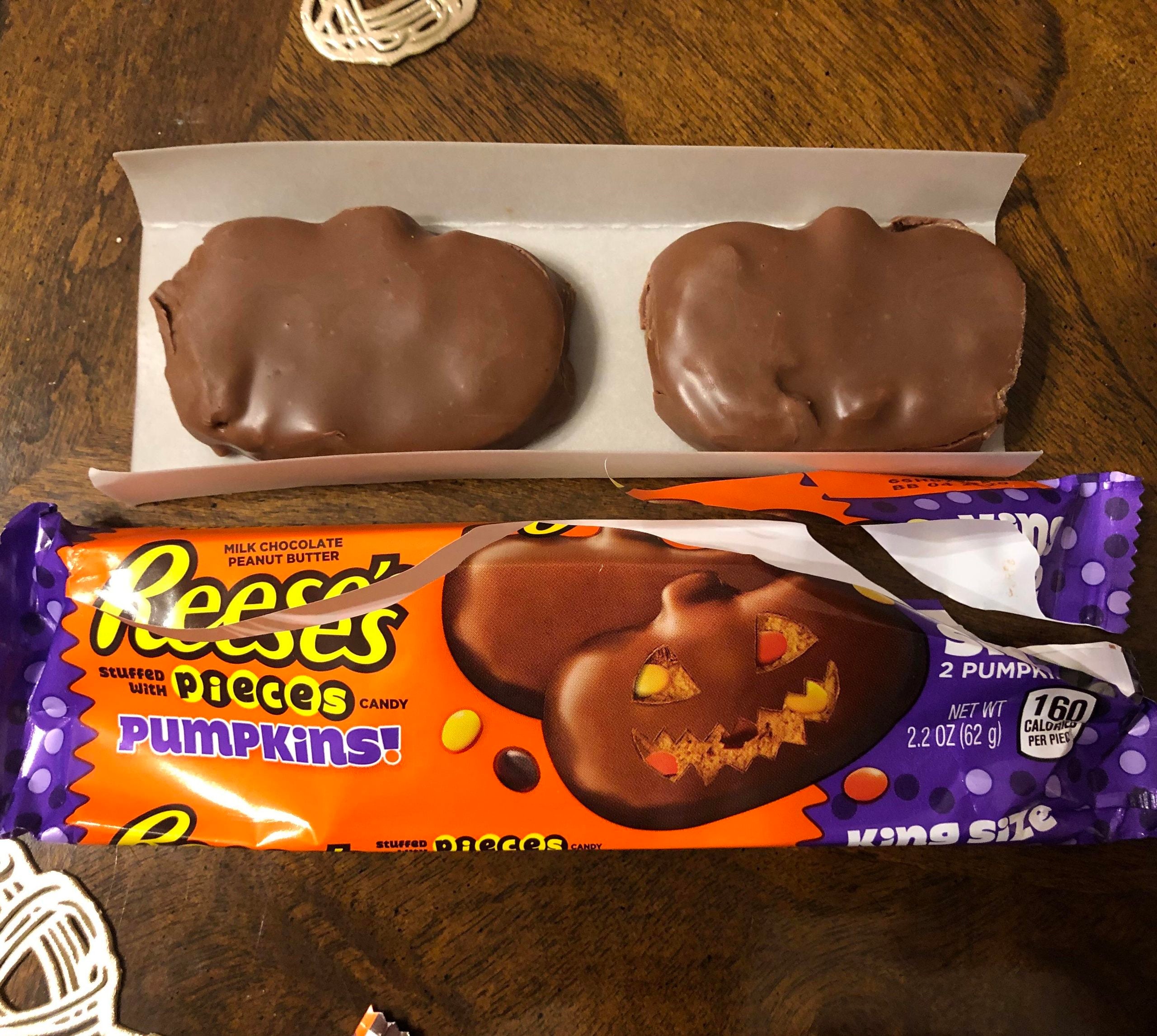 Two chocolate-covered Reese’s Pieces pumpkins in an open package resting above their candy wrapper.
