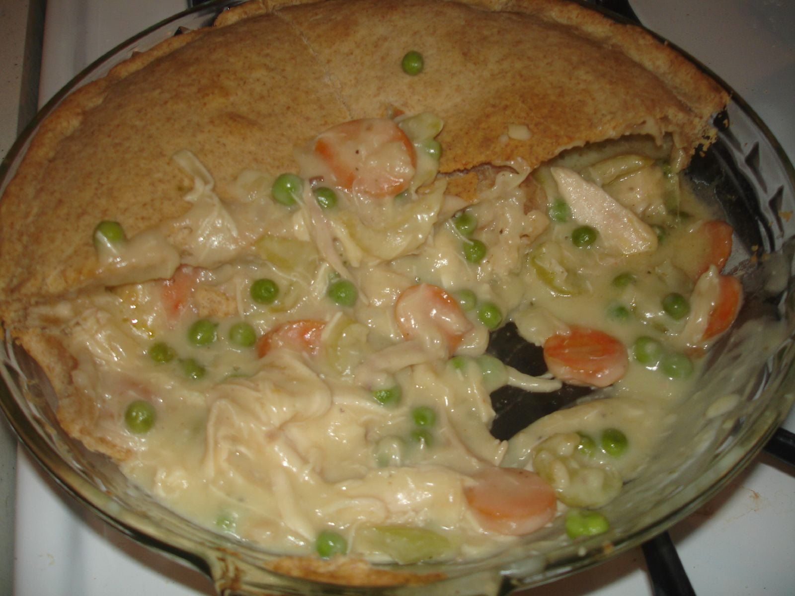 A partially served chicken pot pie with a golden crust, showing creamy filling of chicken, peas, carrots, and celery in a glass dish.
