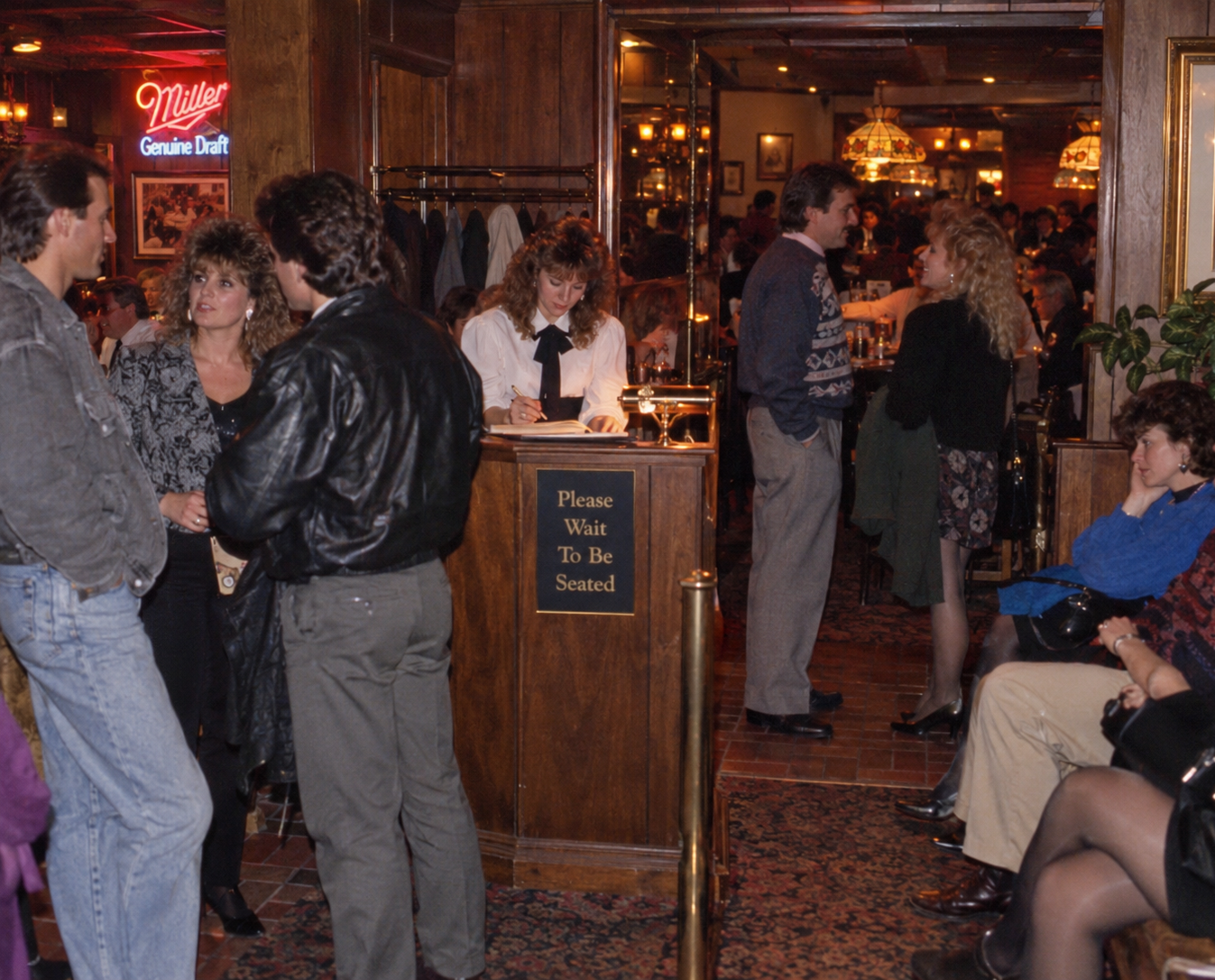 A hostess stands at a wooden podium labeled “Please Wait to Be Seated” while guests wait in a dimly lit restaurant.