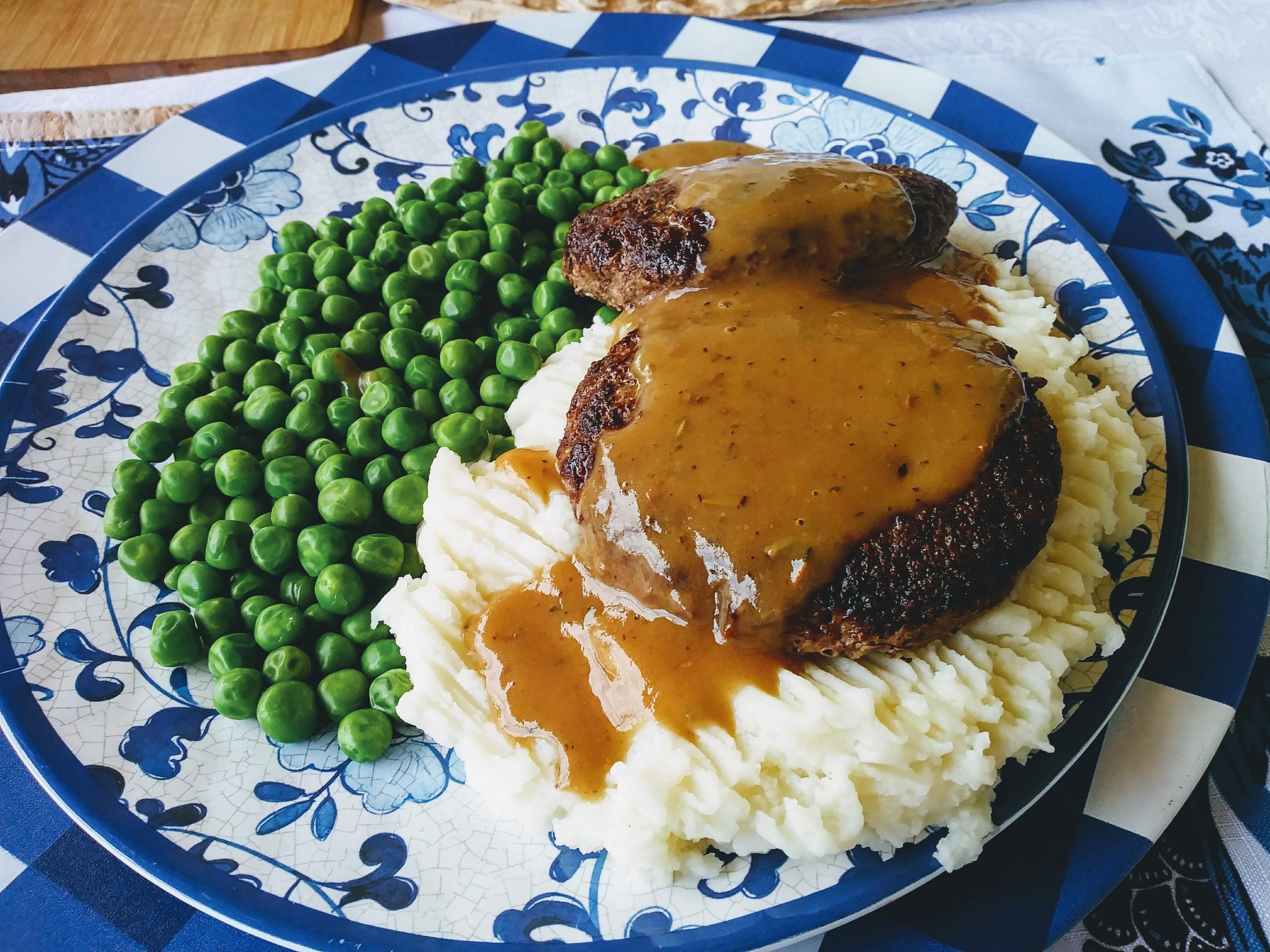 A plate with mashed potatoes topped with brown gravy and meatloaf, served with a side of green peas on a blue and white patterned plate.