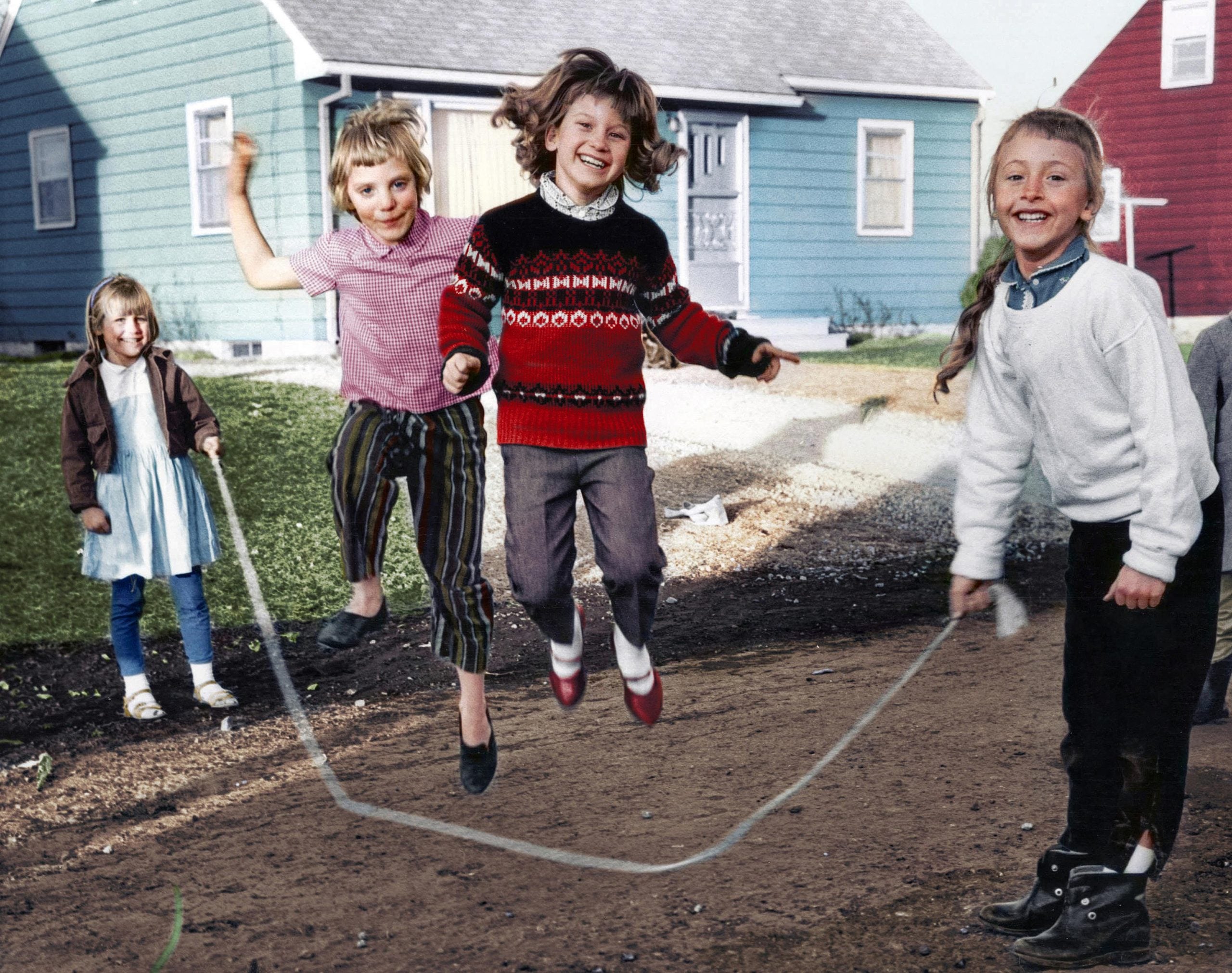 Four children jump rope on a dirt path in front of two houses.