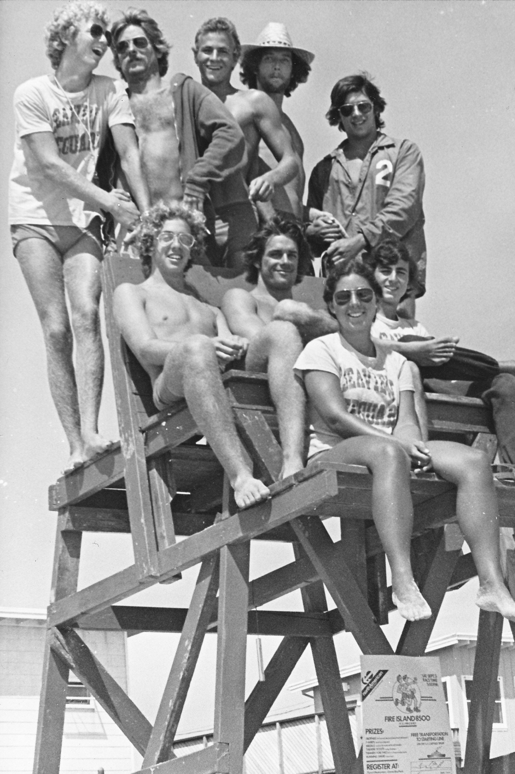 A group of nine young adults sitting and standing on a tall wooden lifeguard chair, with a race registration sign posted on the base.