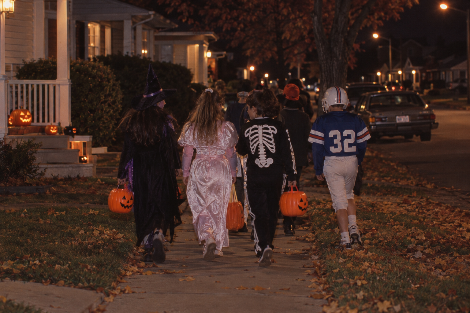 A group of children in Halloween costumes walk down a suburban sidewalk at night carrying pumpkin-shaped candy buckets.