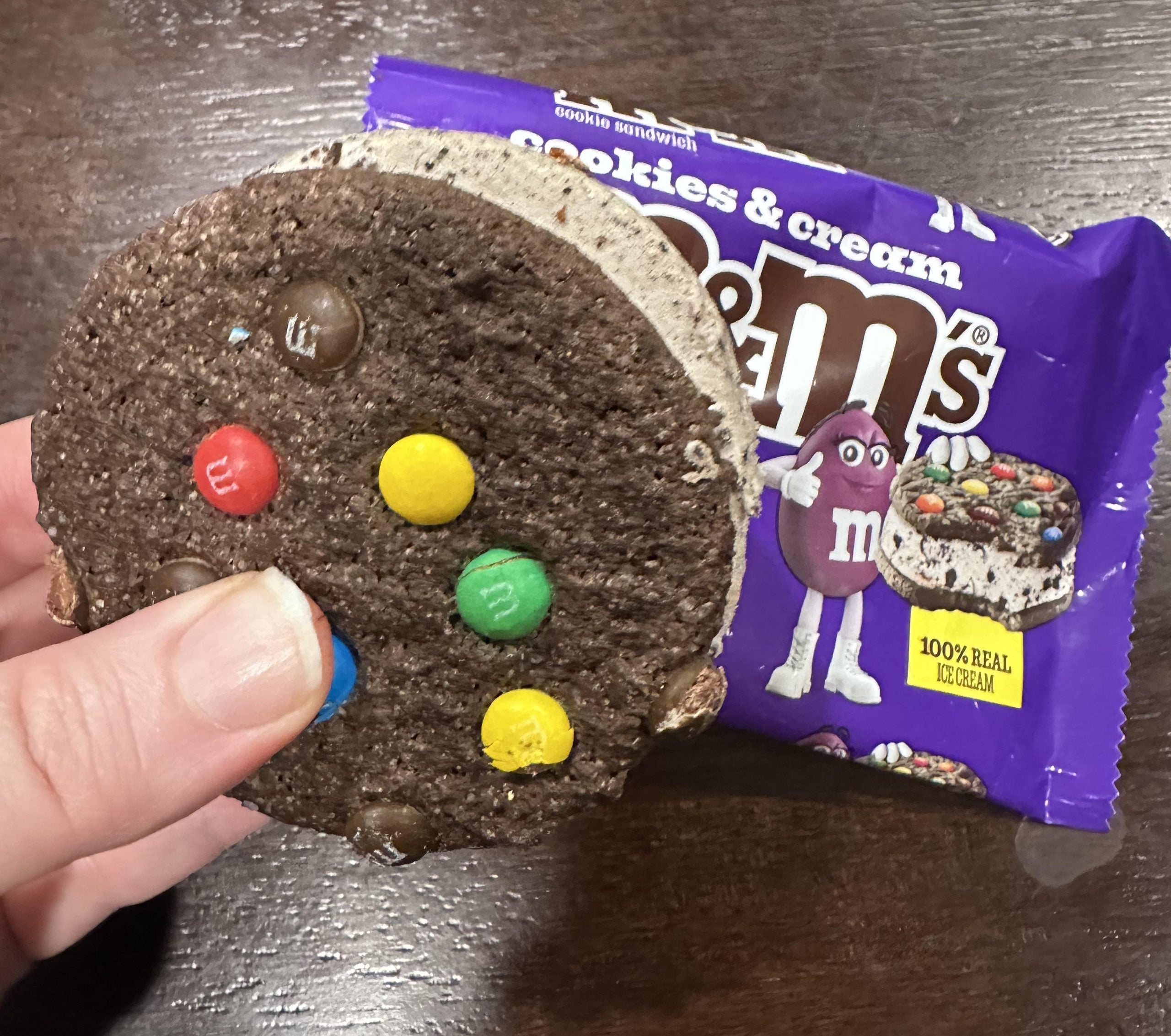 A hand holds a cookies and cream M&M’s ice cream sandwich next to its purple wrapper.