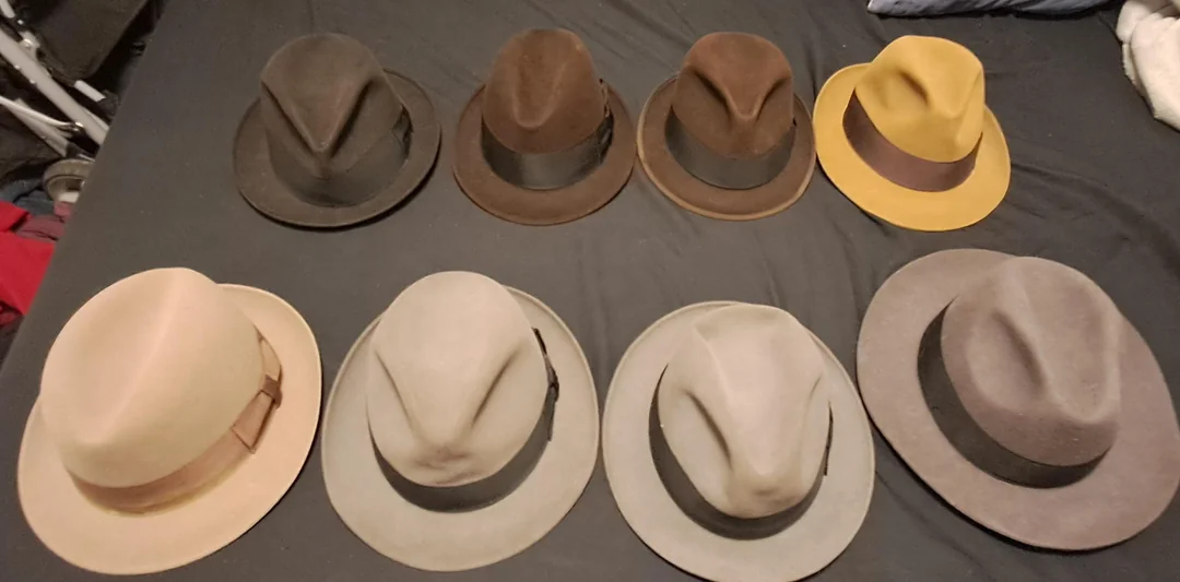 Nine fedora hats in various colors, including black, brown, yellow, tan, and gray, are arranged in two rows on a dark surface. Each hat has a distinct band and crown shape.