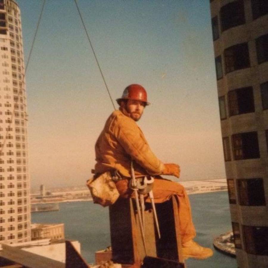 A construction worker in a hard hat and protective gear sits on a steel beam high above a city and harbor.