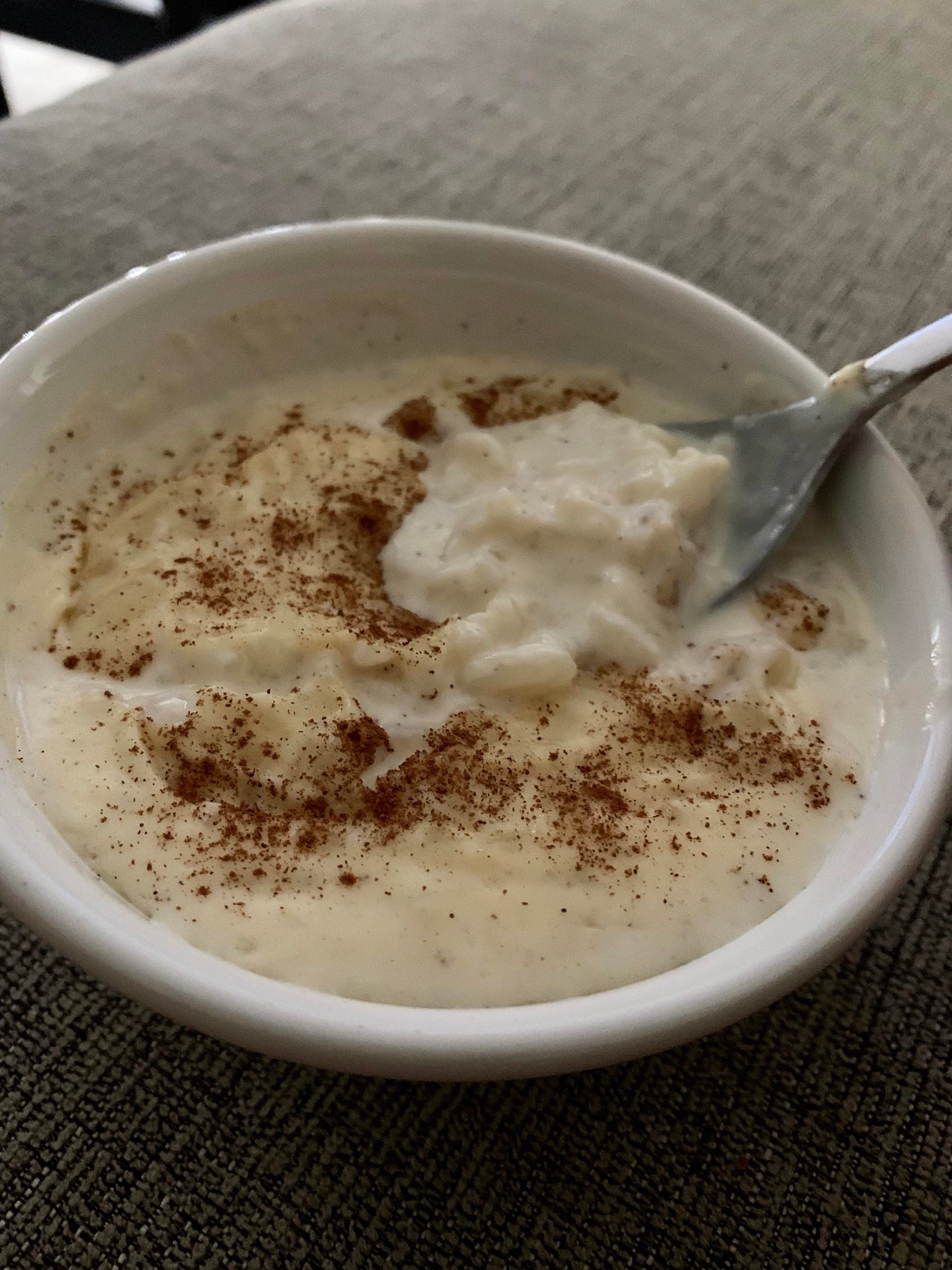 A bowl of rice pudding topped with a sprinkle of cinnamon, with a spoon resting inside, placed on a textured fabric surface.