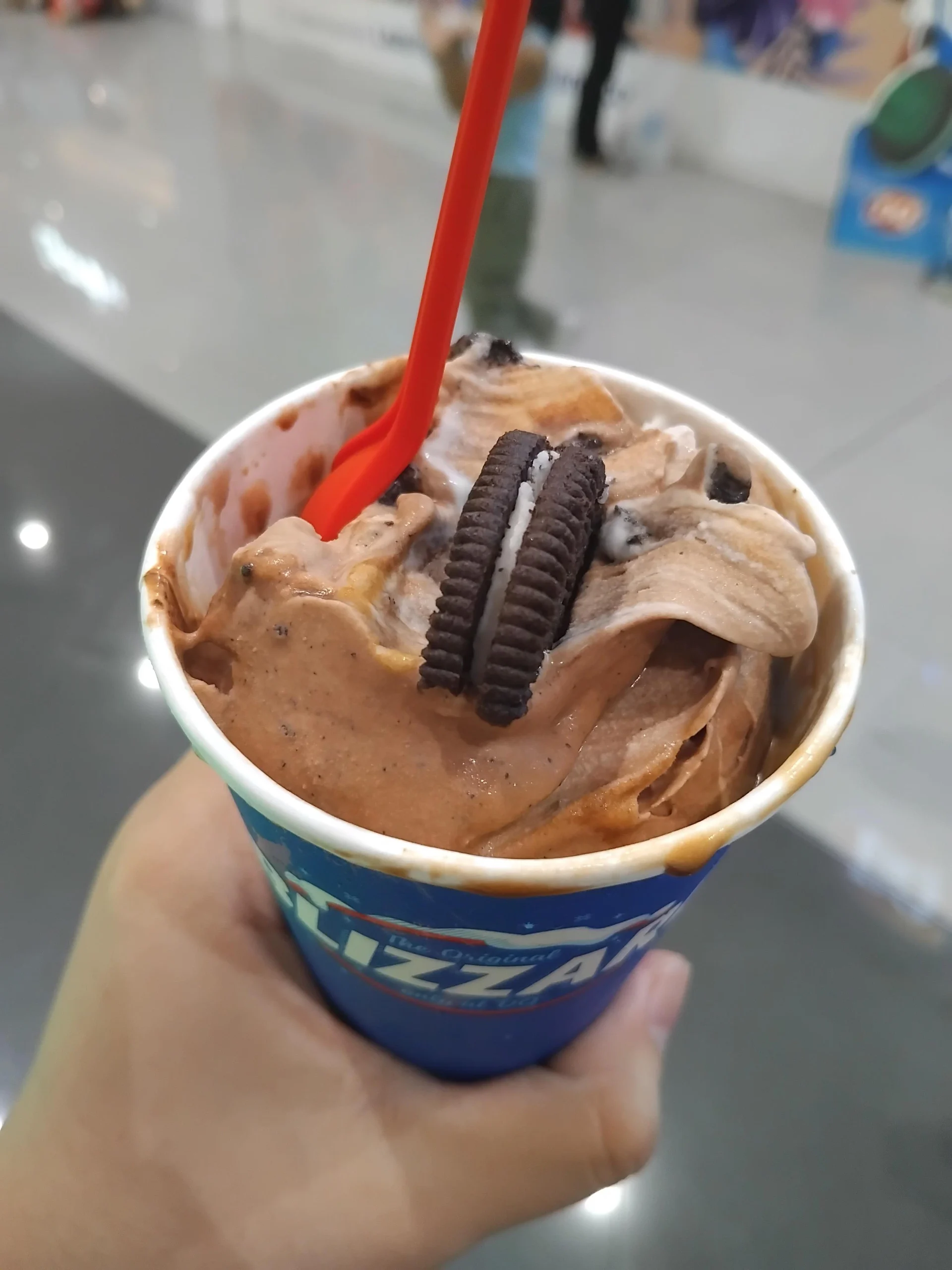 A hand holding a Dairy Queen Blizzard cup filled with chocolate ice cream topped with an Oreo cookie and a red plastic spoon.