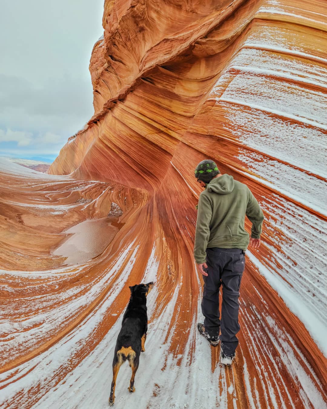 A person in a hooded sweatshirt and a black dog walking through a snow-dusted, orange sandstone rock formation.
