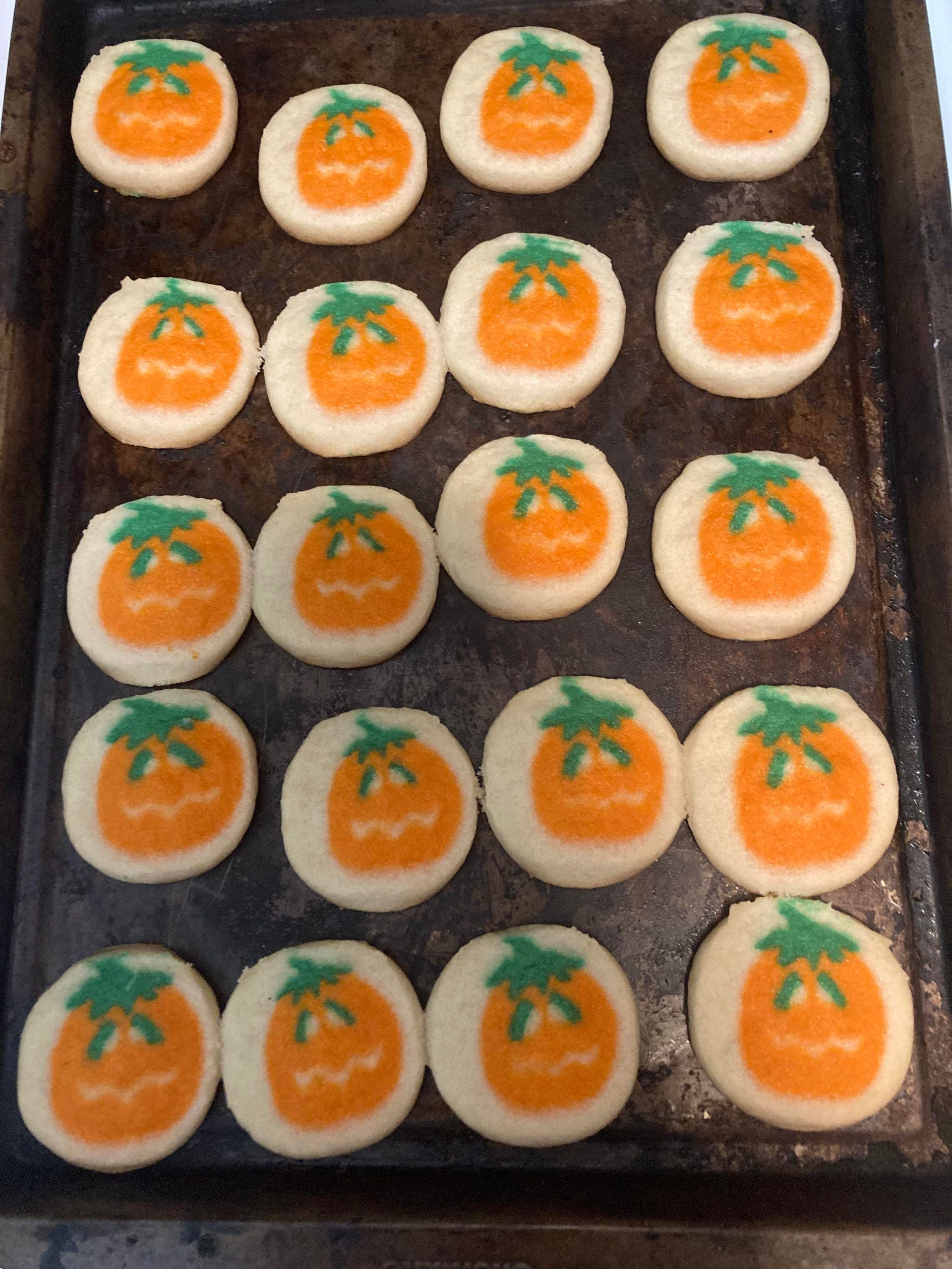 A baking sheet with twenty round sugar cookies decorated to look like orange pumpkins with green stems.