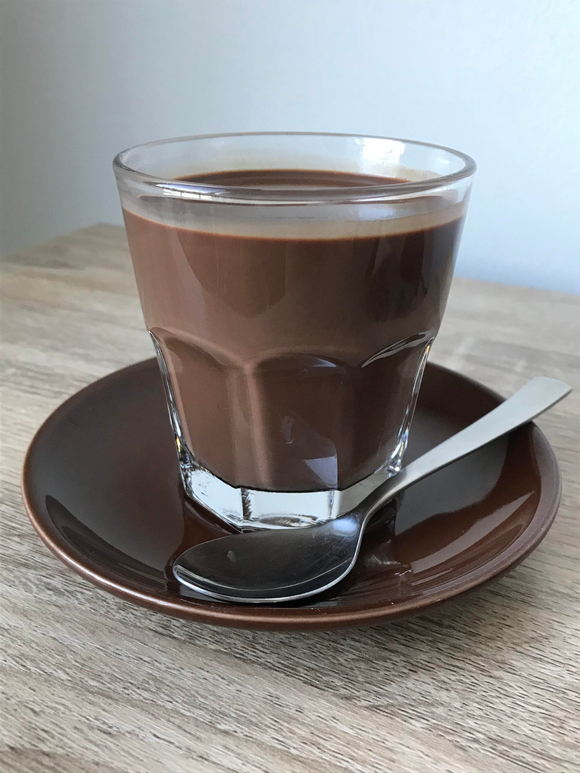A glass of chocolate milk in a brown saucer with a silver spoon.