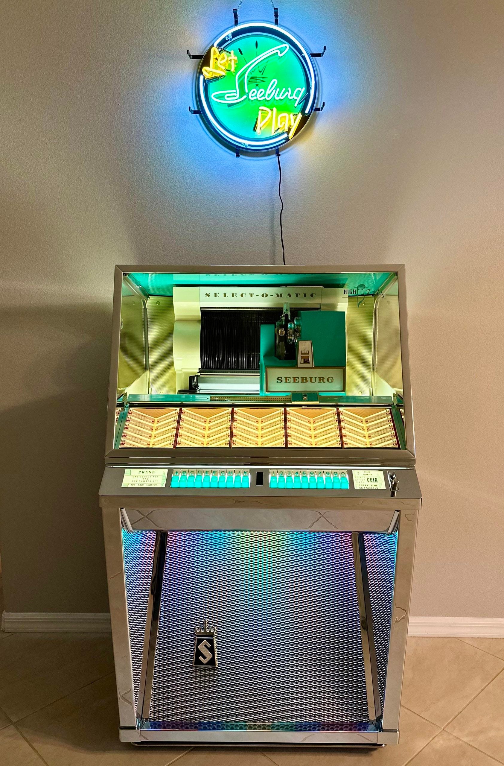 A vintage Seeburg jukebox with colorful lights stands against a beige wall beneath a glowing blue and yellow neon sign that reads “Let Seeburg Play.”