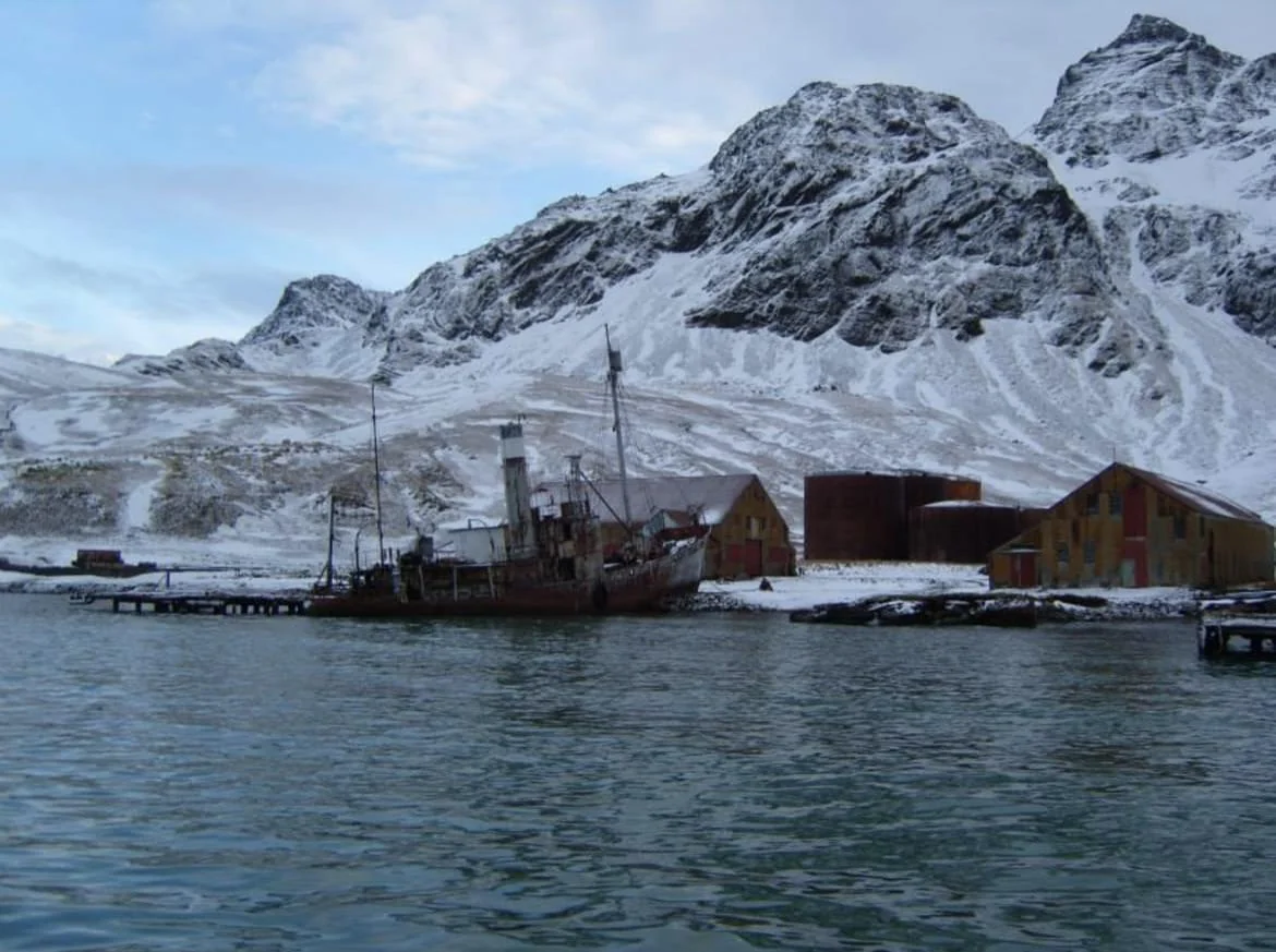 An old, rusted ship docked at a snowy whaling station with large fuel tanks, set against a backdrop of steep, snow-covered mountains.