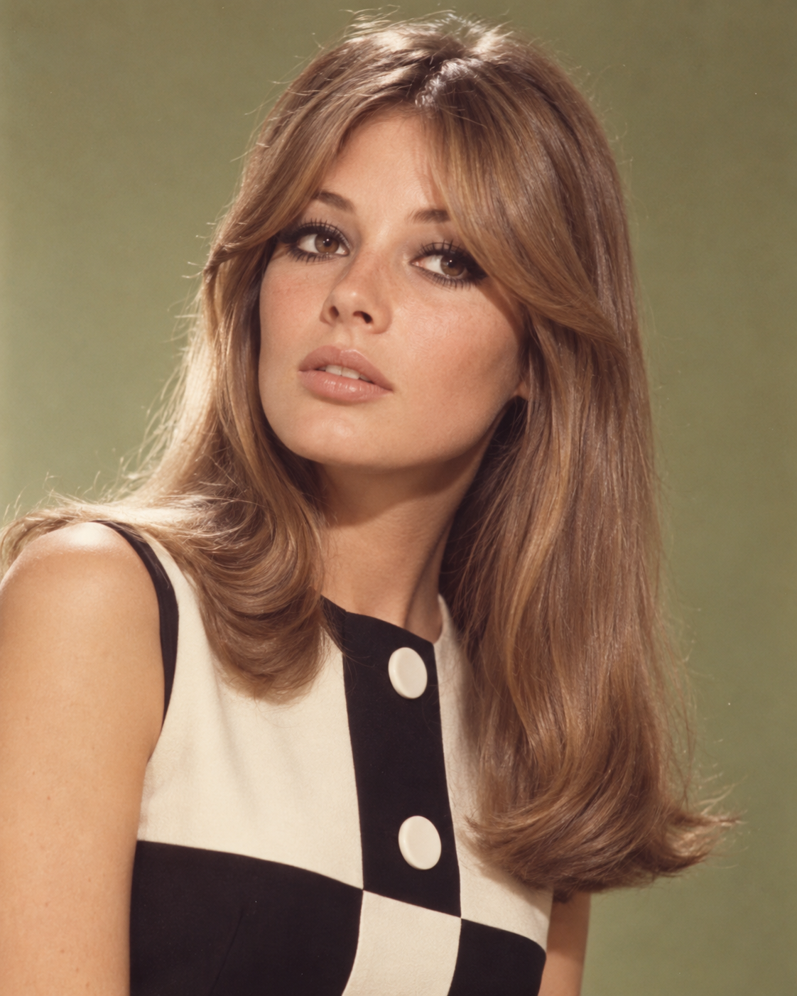 A portrait of a woman with long, layered brown hair wearing a white and black sleeveless dress with large buttons.