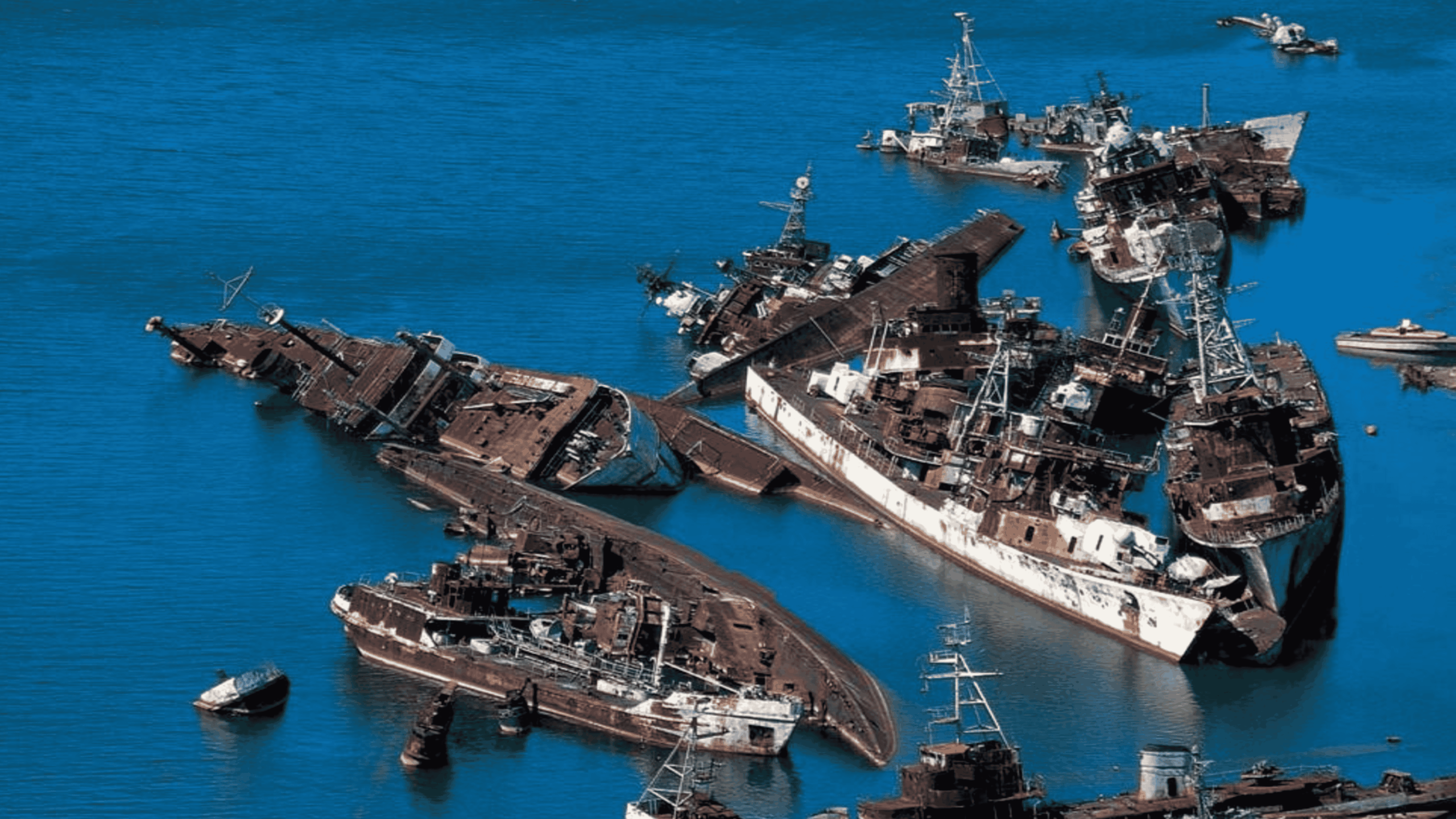 An aerial view of several rusted, partially submerged shipwrecks grouped together in blue water.