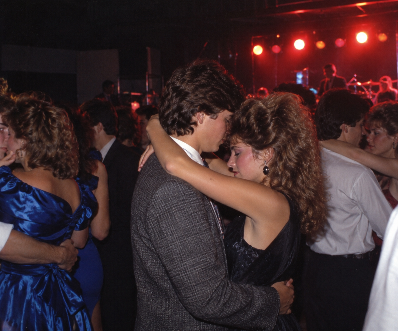 A young couple slow dances at a crowded party with stage lights in the background.