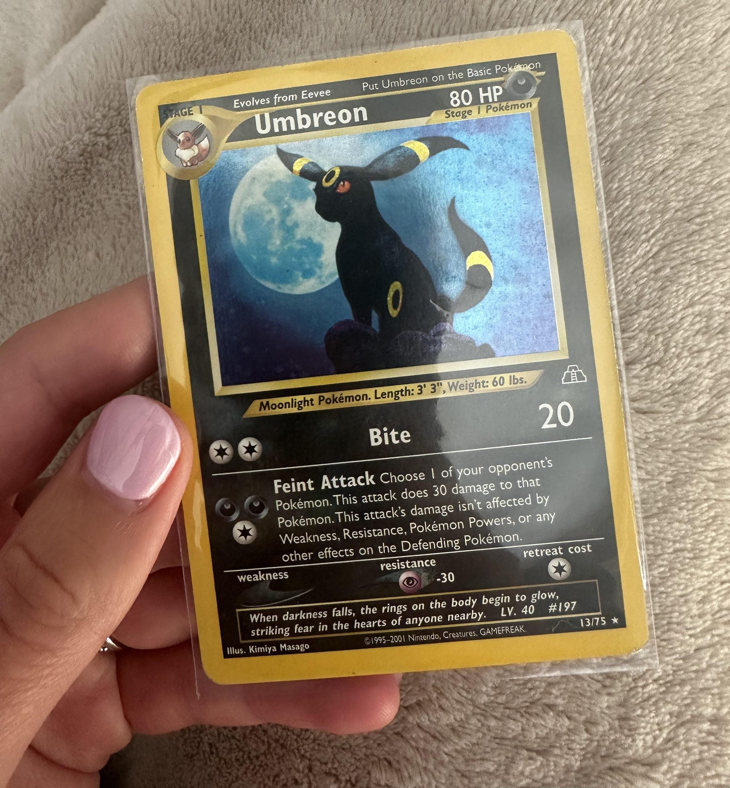 A hand with pink-painted nails holds a holographic Umbreon Pokémon card in a plastic sleeve over a beige textured surface. The card displays Umbreon with moonlit details and its attack information.