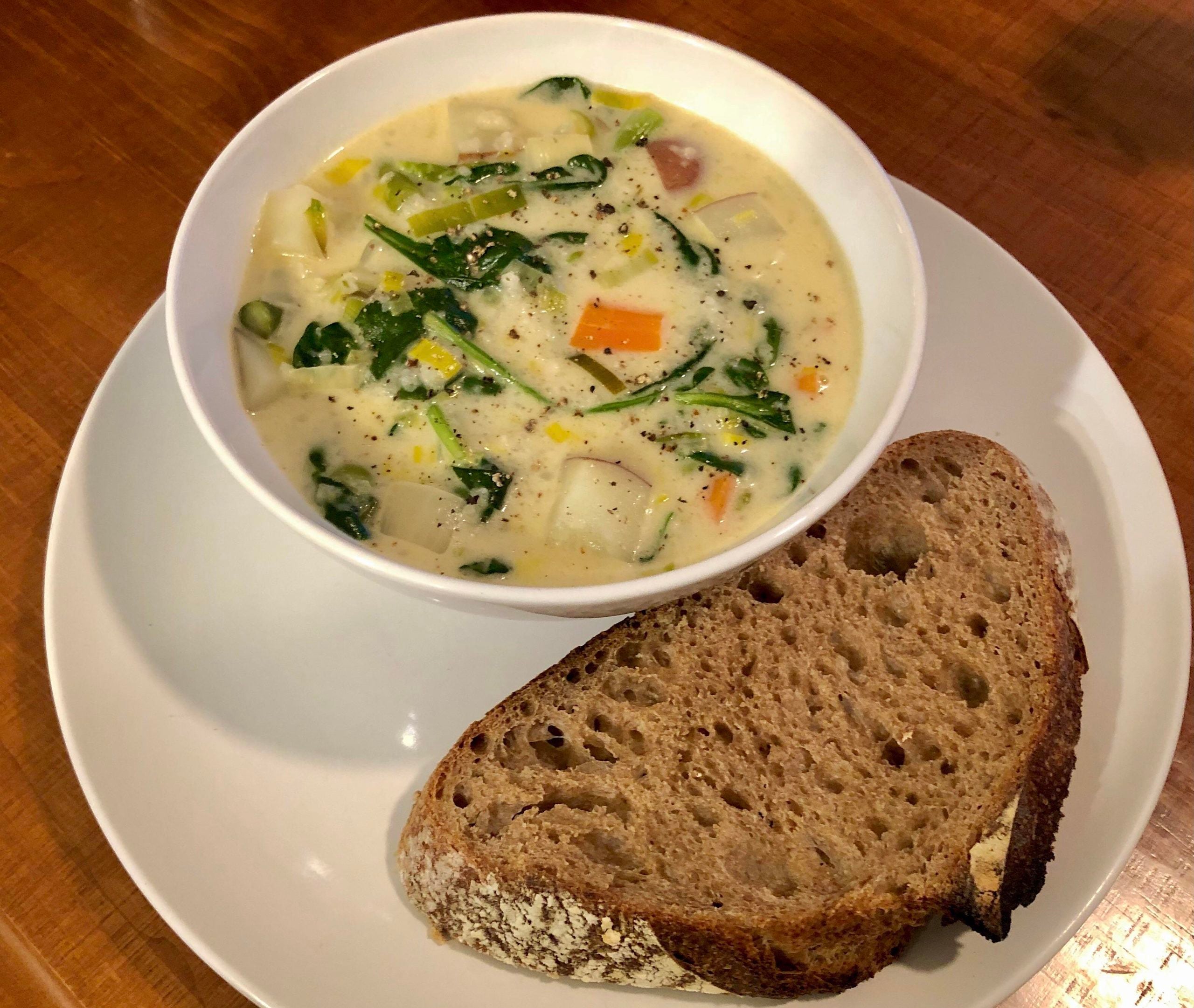 A bowl of creamy vegetable soup with greens, potatoes, and carrots, served on a white plate with a slice of rustic brown bread.