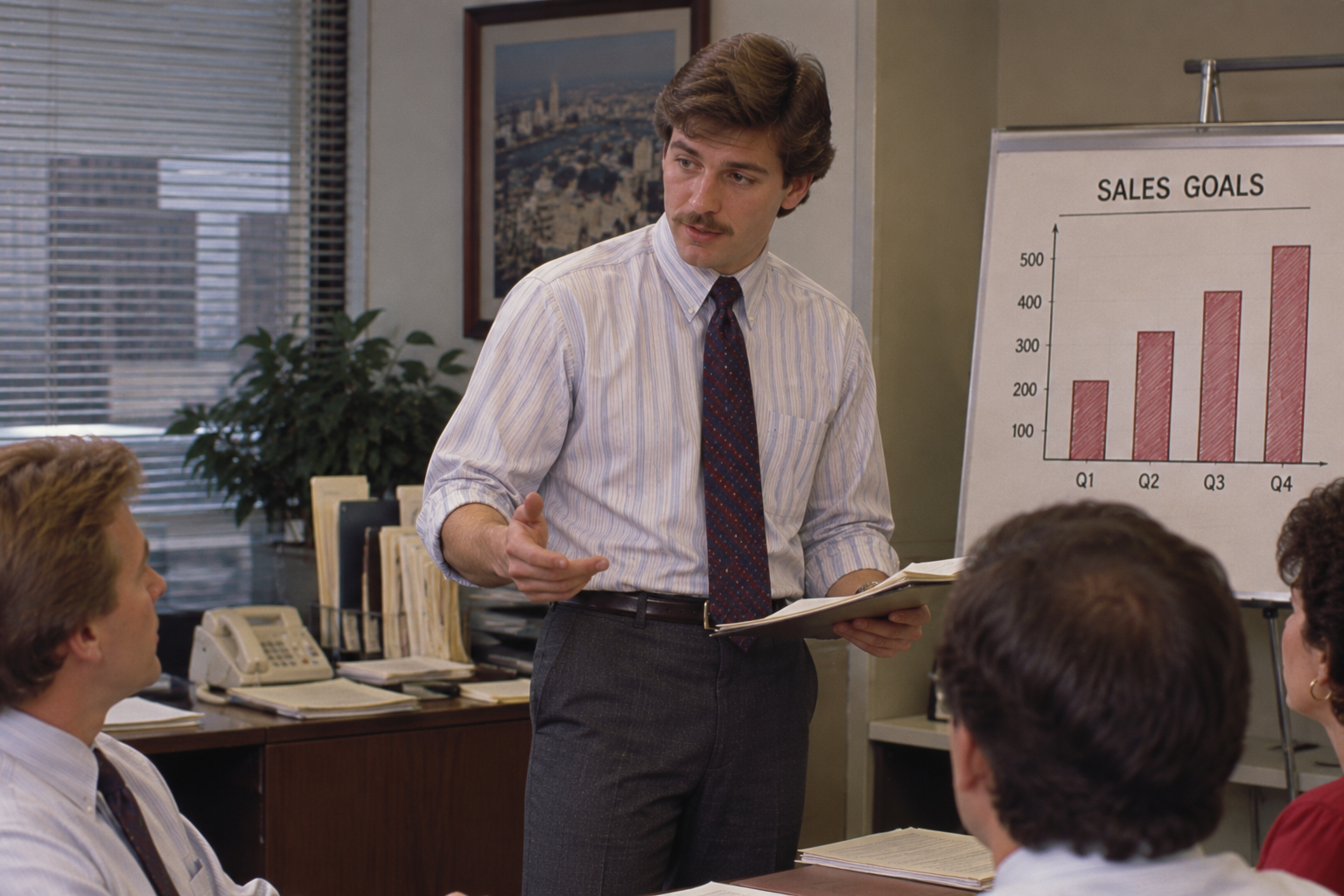 A man with a mustache stands in an office giving a presentation to colleagues in front of a whiteboard displaying a bar chart labeled “Sales Goals.”