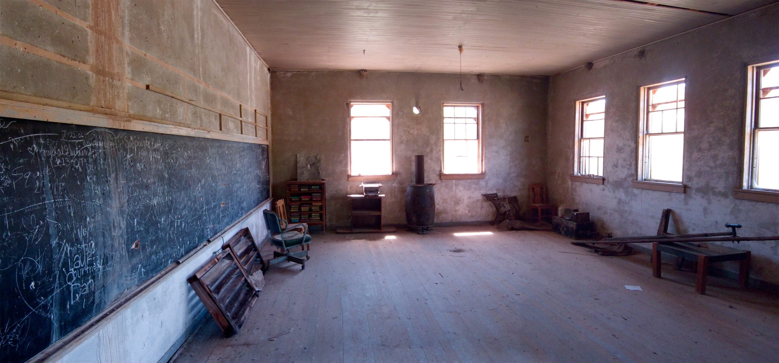 The interior of a dilapidated one-room schoolhouse with a large blackboard covered in chalk writing, several windows, and sparse, abandoned furniture.