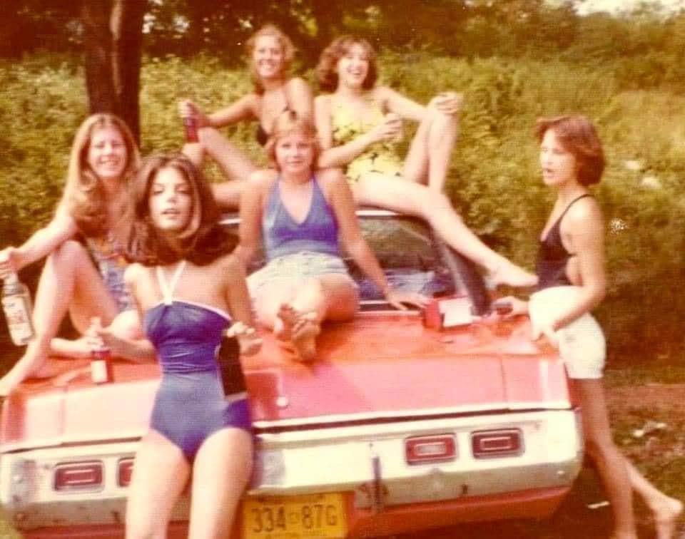 Six young women posing in swimwear on and around the back of a red car.