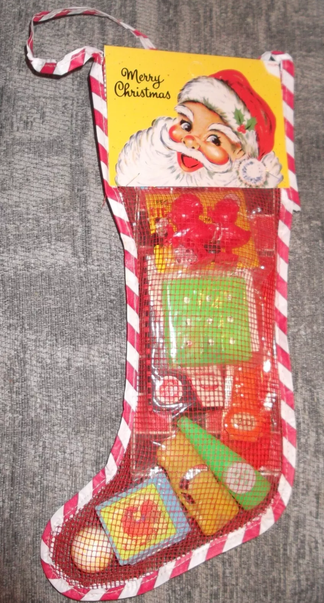 A Christmas stocking filled with small plastic toys, featuring a cardboard topper illustration of Santa Claus and the text “Merry Christmas.”