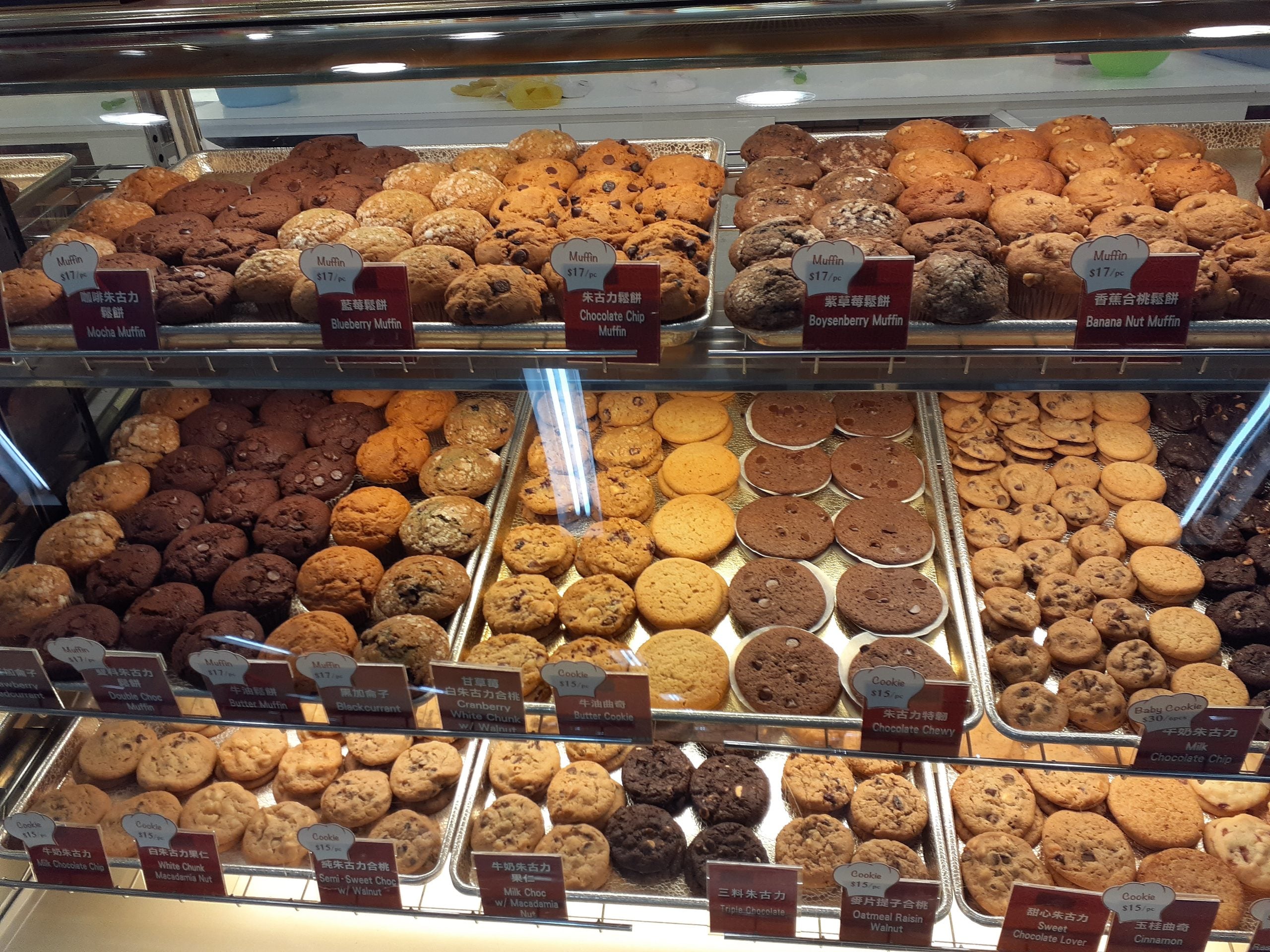 A glass display case filled with rows of assorted muffins and cookies, each with a small label indicating its flavor and price.