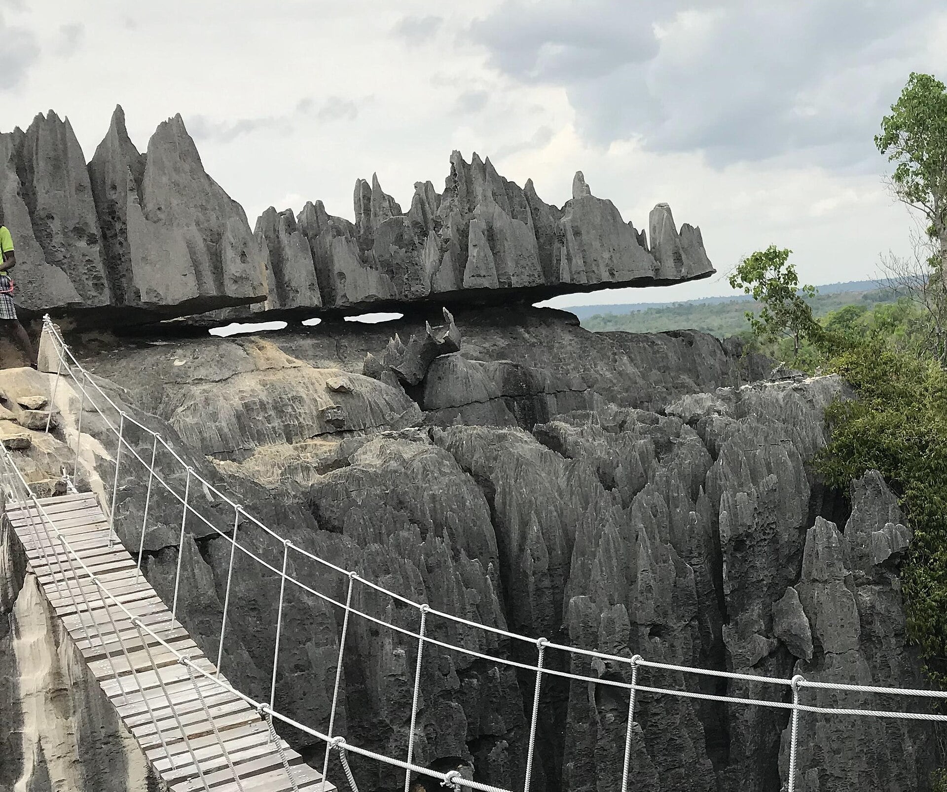 A rope bridge leads to a jagged, grey rock formation set against a cloudy sky.