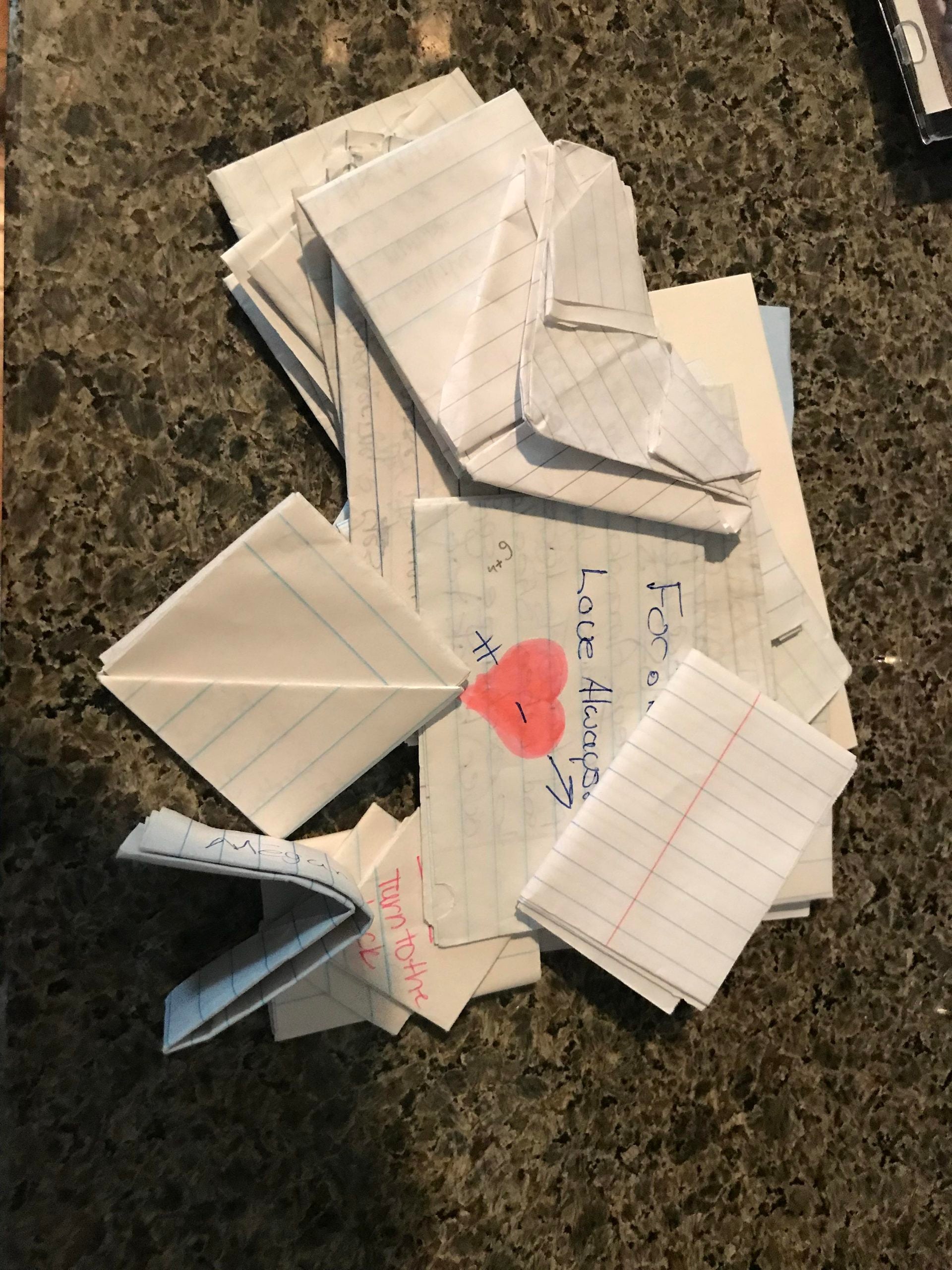 A pile of folded note-passing papers made from notebook paper, one showing a hand-drawn heart and the text “For you, Love Always.”